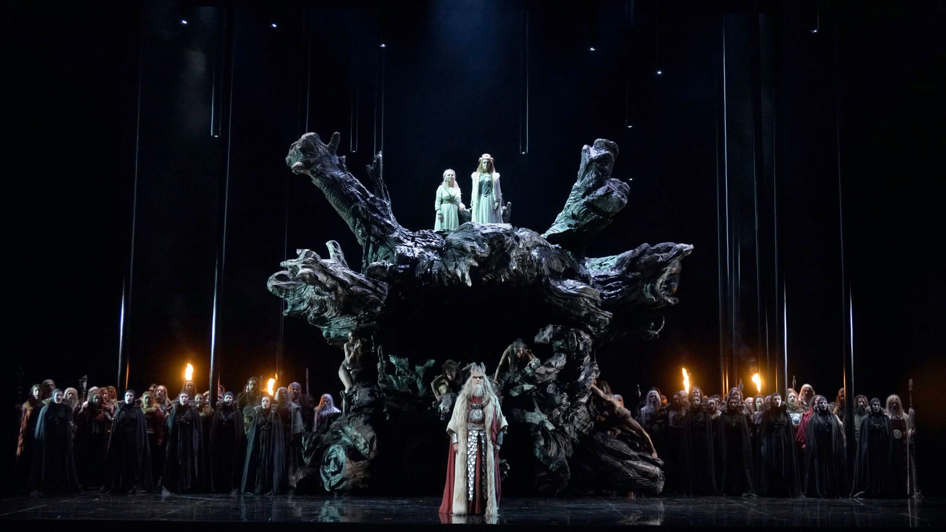 Enorme radice d'albero annerita al centro del palco con due donne in abiti chiari in cima e il coro schierato sotto – Norma al Teatro Real, Madrid, Spagna, 2016 – scenografia d'opera