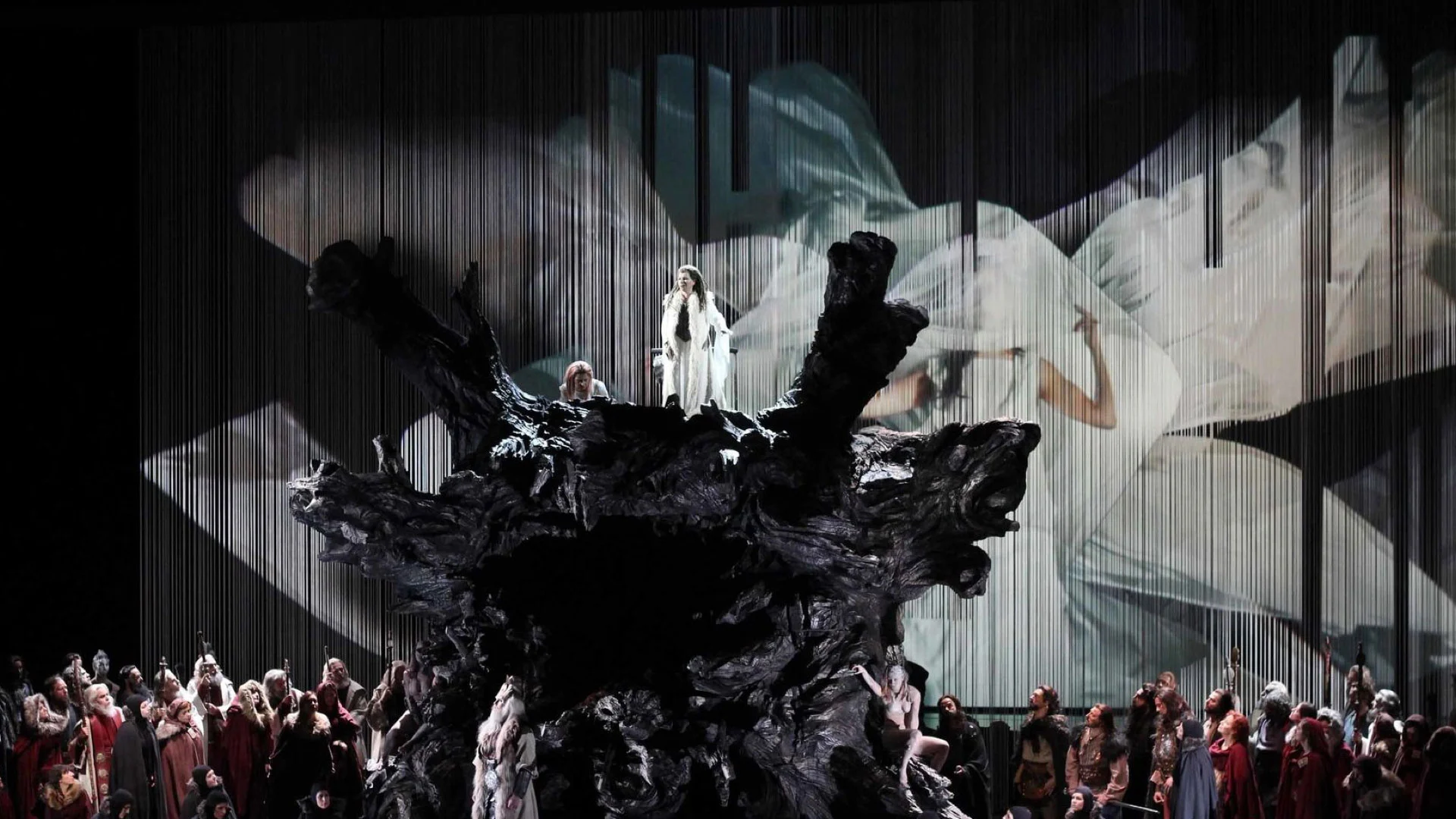 Scenografia di una radice d'albero scura e scolpita con una figura in bianco in cima, davanti alla proiezione di un drappo bianco gonfio – Norma al Teatro Real, Madrid, Spagna, 2016 – scenografia d'opera