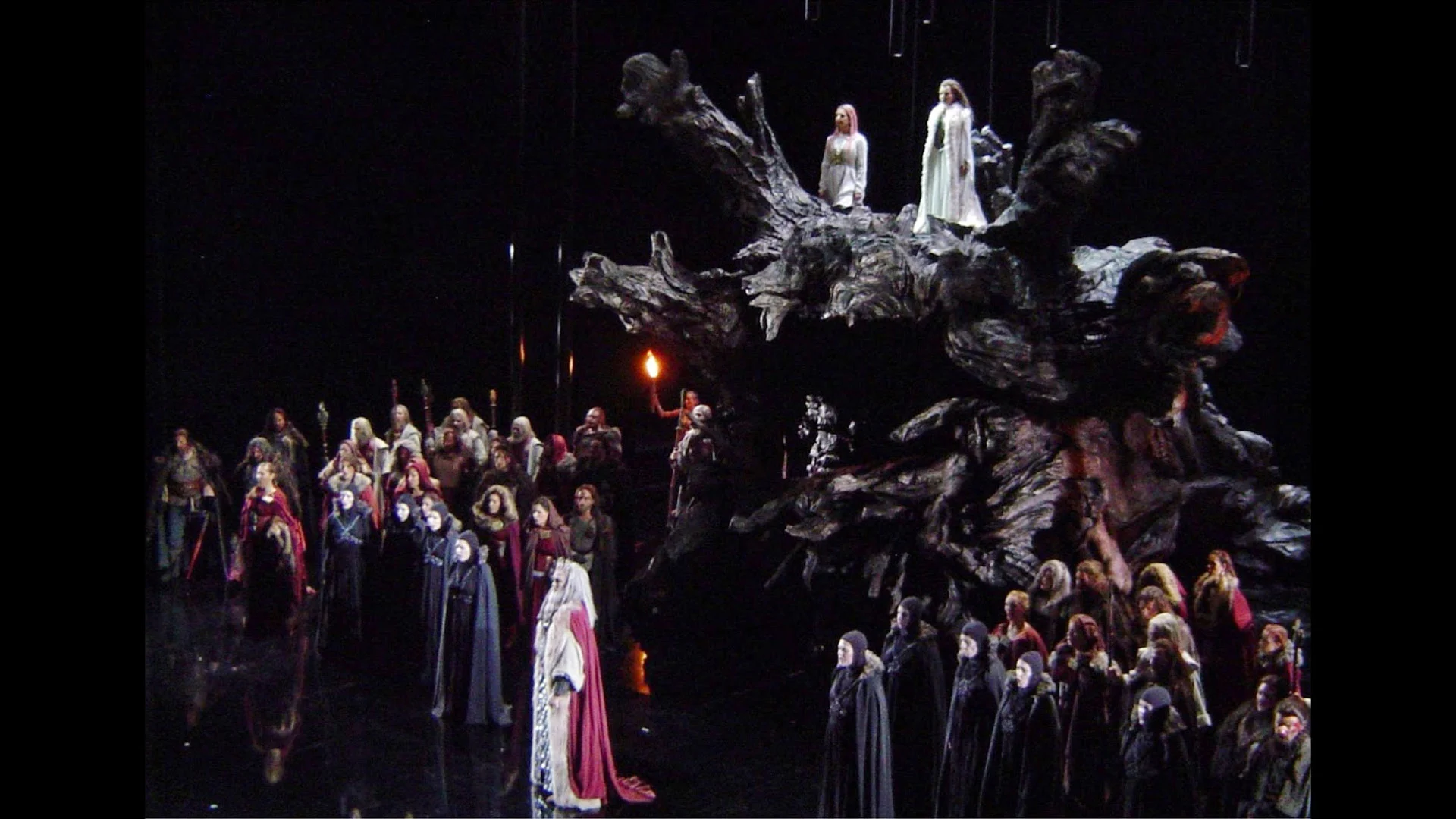 Vista laterale della scenografia a radice d'albero con due figure in cima e il coro incappucciato con le torce – Norma al Teatro Real, Madrid, Spagna, 2016 – scenografia d'opera