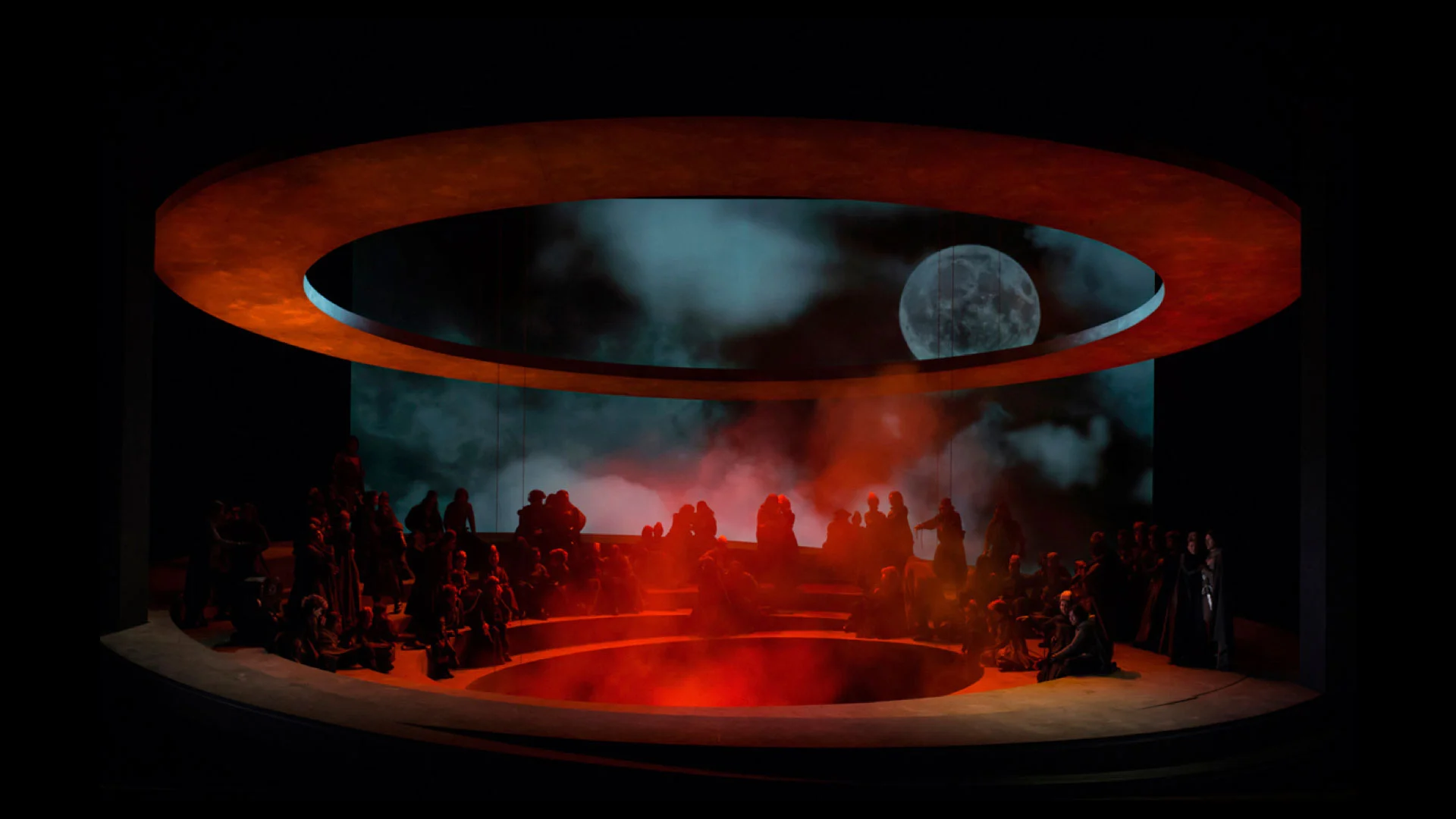 Palco circolare a gradoni in luce rossa e fumo sotto un anello arancione, con luna e nuvole proiettate – Otello al Teatro Carlo Felice, Genova, Italia, 2014 – scenografia d'opera