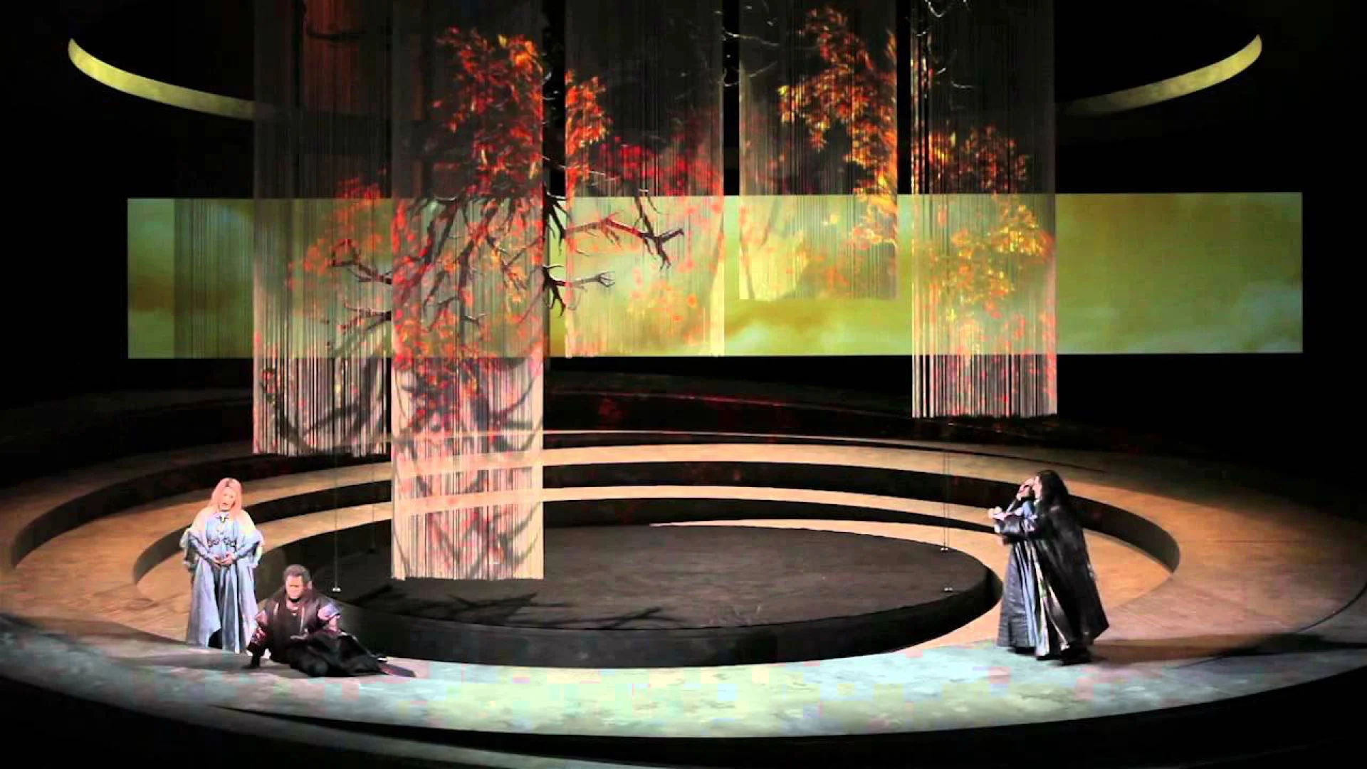 Tre cantanti sul palco circolare sotto tende a fili che mostrano un albero autunnale in rosso e oro – Otello al Teatro Carlo Felice, Genova, Italia, 2014 – scenografia d'opera