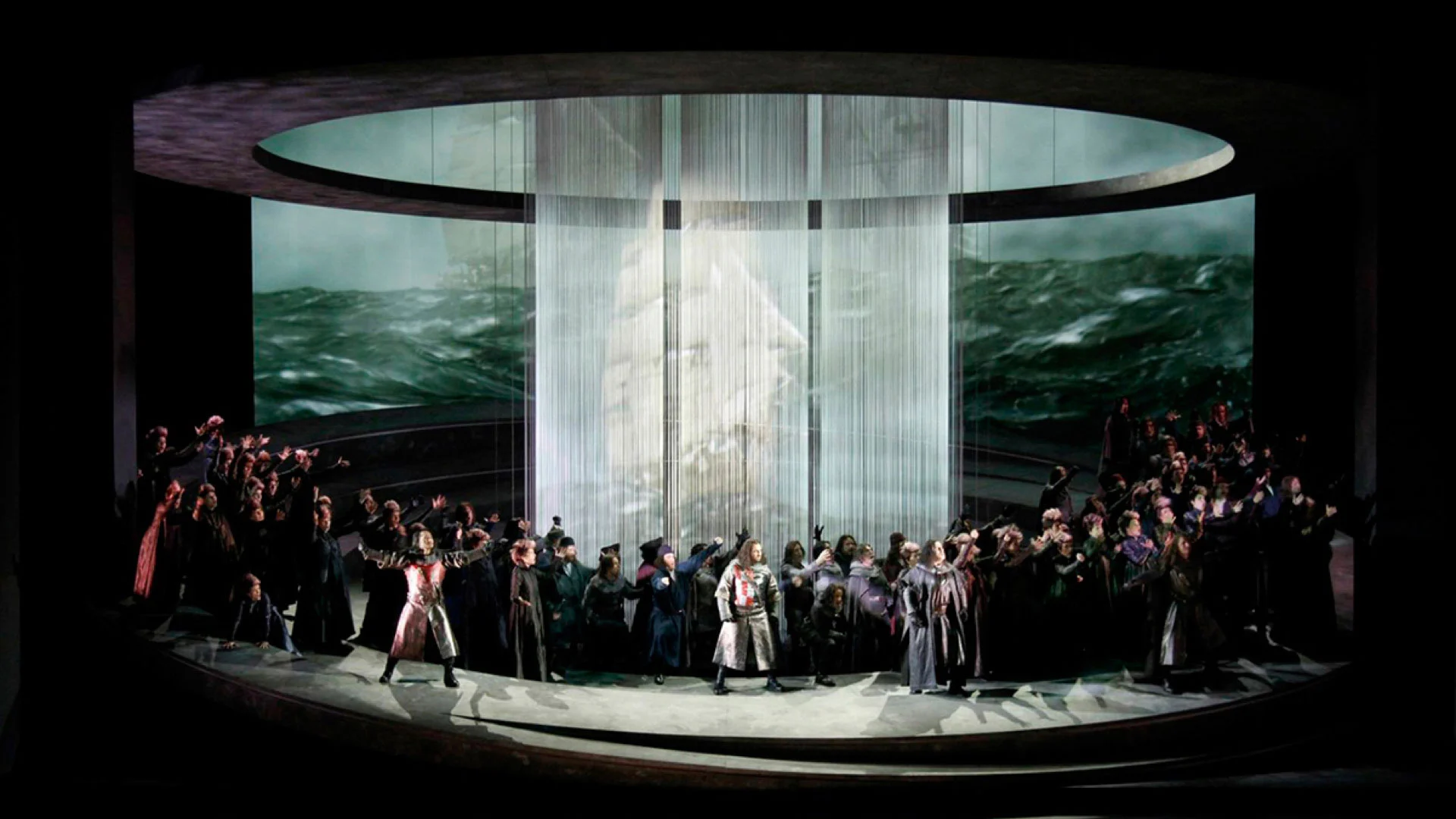 Coro sul palco circolare davanti a tende a fili con la proiezione di un veliero in un mare in tempesta – Otello al Teatro Carlo Felice, Genova, Italia, 2014 – scenografia d'opera