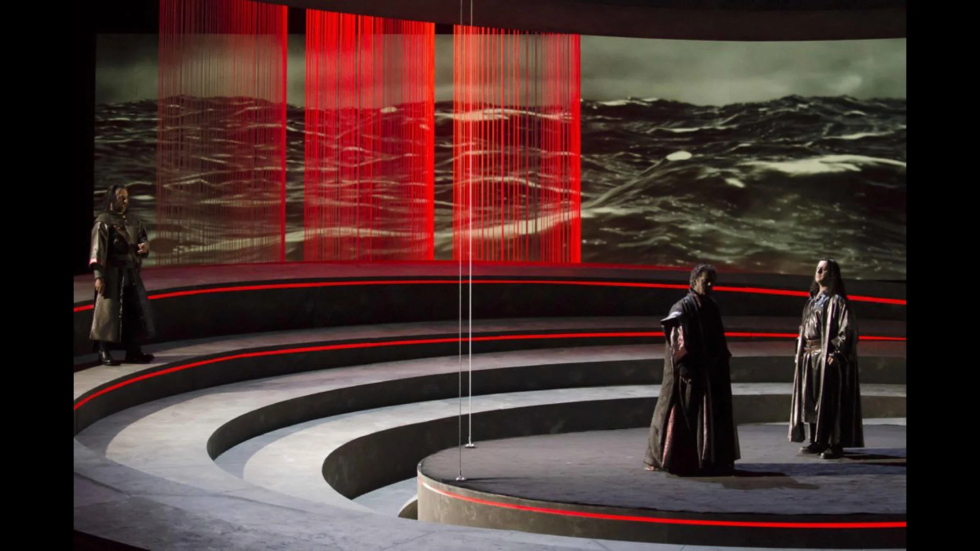 Tre artisti sui gradoni curvi davanti alla proiezione di un mare in tempesta attraversata da tende a fili rosse – Otello al Teatro Carlo Felice, Genova, Italia, 2014 – scenografia d'opera