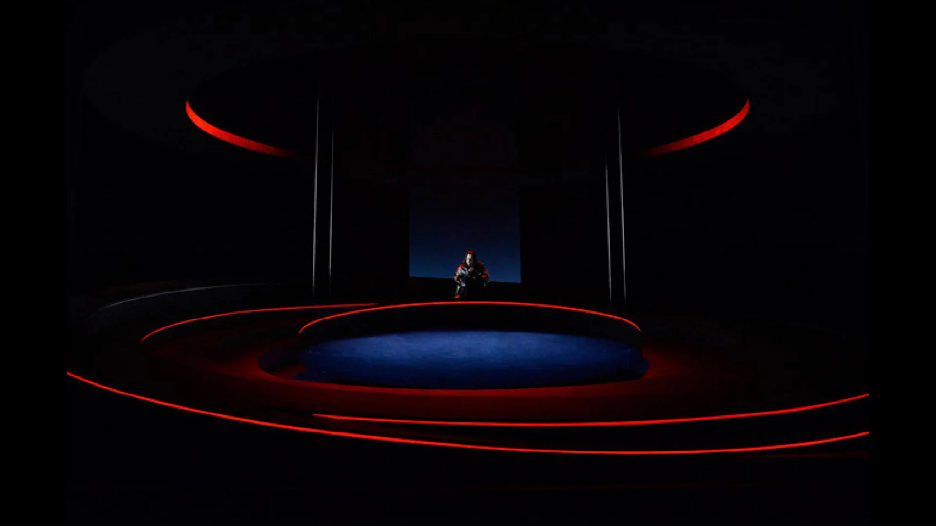 Palco buio con una figura solitaria seduta al centro, delineata da anelli di luce rossa sui gradoni – Otello al Teatro Carlo Felice, Genova, Italia, 2014 – scenografia d'opera