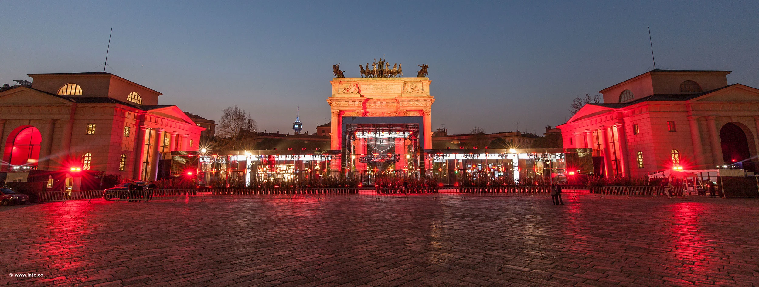 Ampia vista al crepuscolo della piazza e dell'arco, con lunghi padiglioni illuminati di rosso ed edifici ai lati – Cartier The Legendary Thrill, Milano, Italia, 2018 – progettazione di eventi