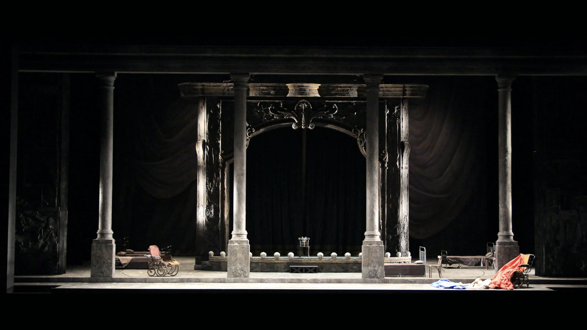 Scenografia con alte colonne che inquadrano un portale teatrale decorato, una carrozzina e un letto sul palco in penombra – Adriana Lecouvreur all'Opéra de Monte-Carlo, Principato di Monaco, 2026 – scenografia d'opera
