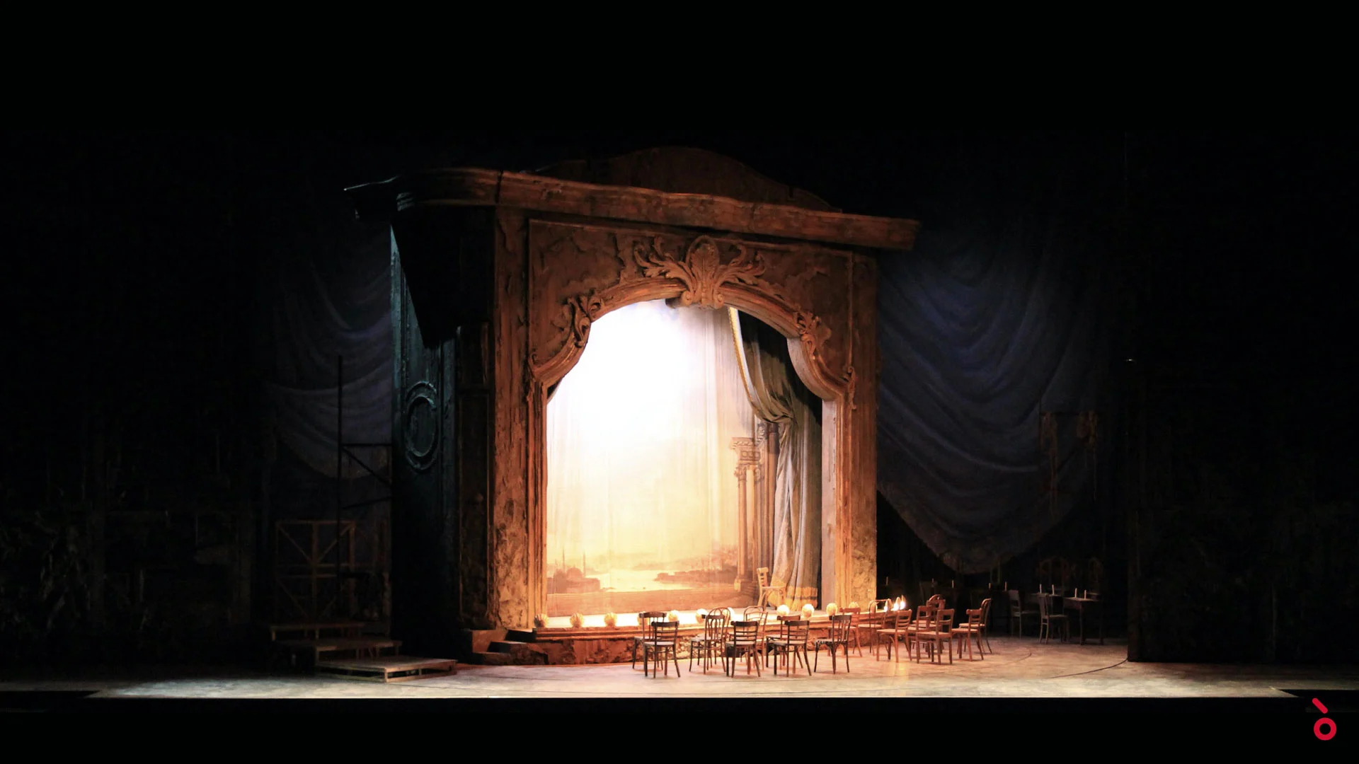 Portale teatrale intagliato illuminato con un fondale dipinto, tavolini e sedie da caffè davanti – Adriana Lecouvreur all'Opéra de Monte-Carlo, Principato di Monaco, 2026 – scenografia d'opera