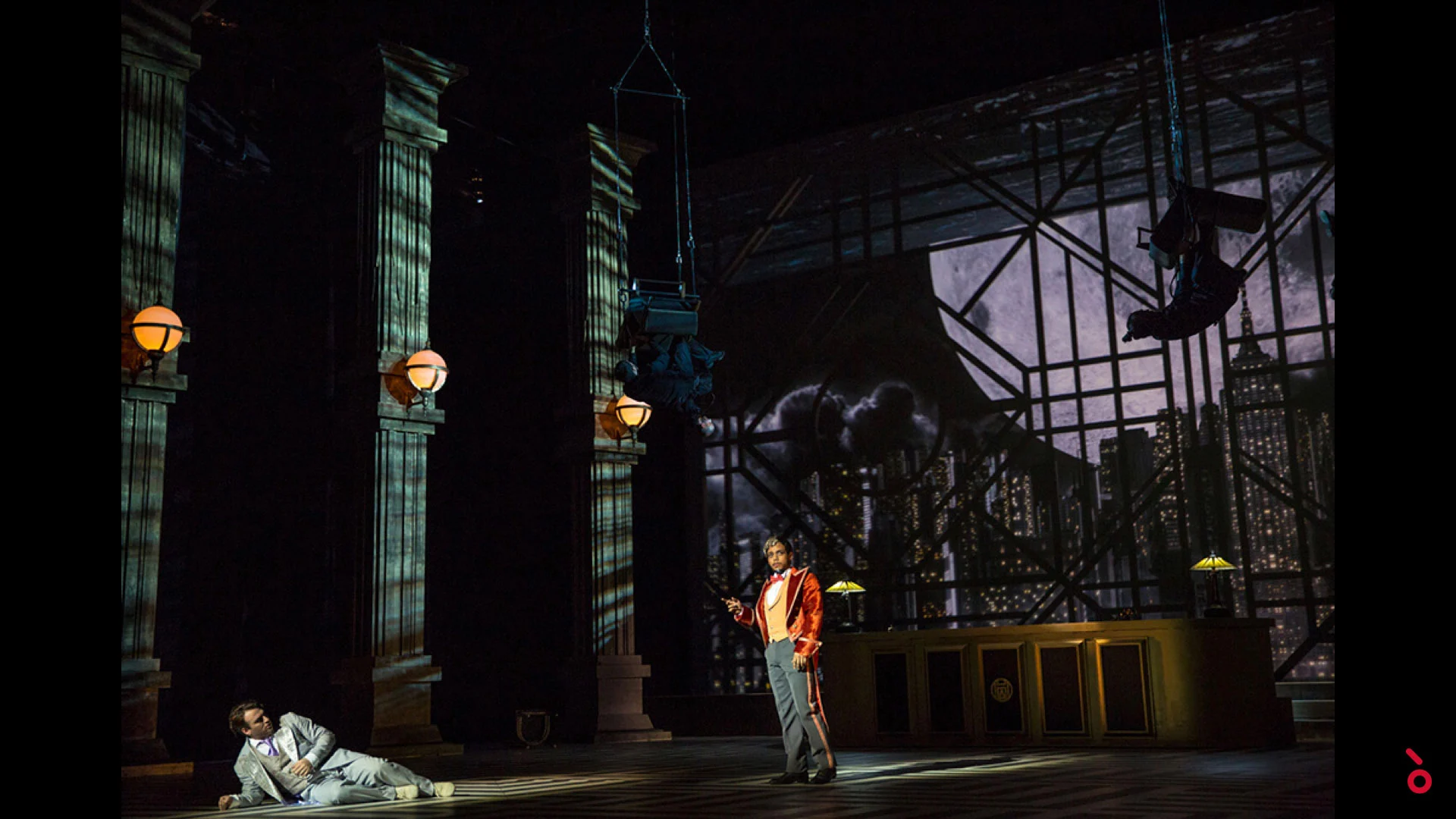 Due artisti in una hall in penombra con alte colonne e lampade a globo, uno skyline notturno alle spalle – The Opera! alla Royal Opera House Muscat, Oman, 2019 – scenografia