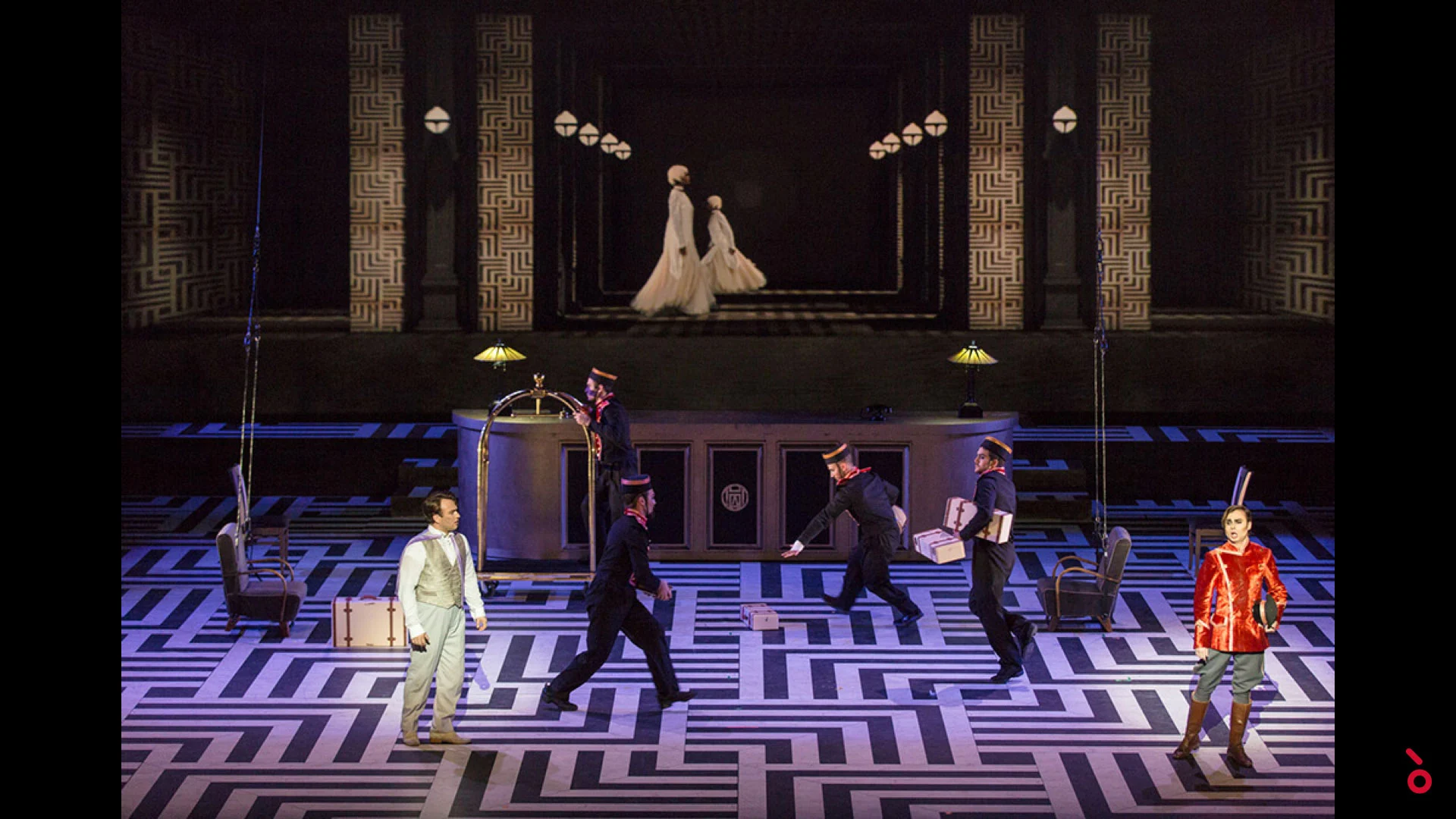 Fattorini che portano bagagli attraverso la scenografia dell'hotel mentre due donne in bianco percorrono una galleria sul fondo – The Opera! alla Royal Opera House Muscat, Oman, 2019 – scenografia