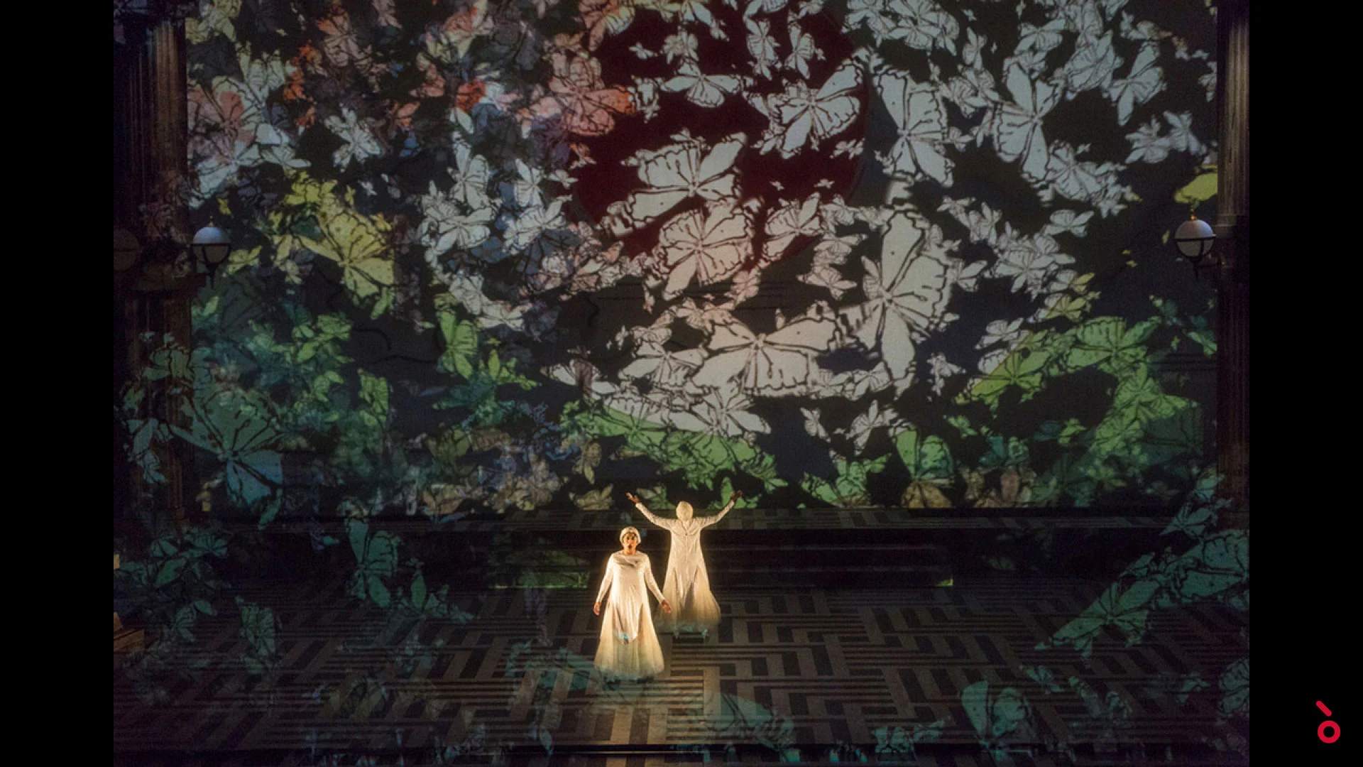 Due donne in bianco su un palco buio coperto da un motivo proiettato di farfalle e foglie – The Opera! alla Royal Opera House Muscat, Oman, 2019 – scenografia