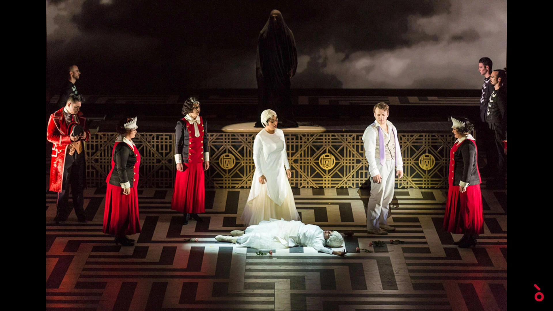 Cast in costumi rossi e bianchi attorno a una figura distesa su un pavimento geometrico, una statua velata sopra – The Opera! alla Royal Opera House Muscat, Oman, 2019 – scenografia