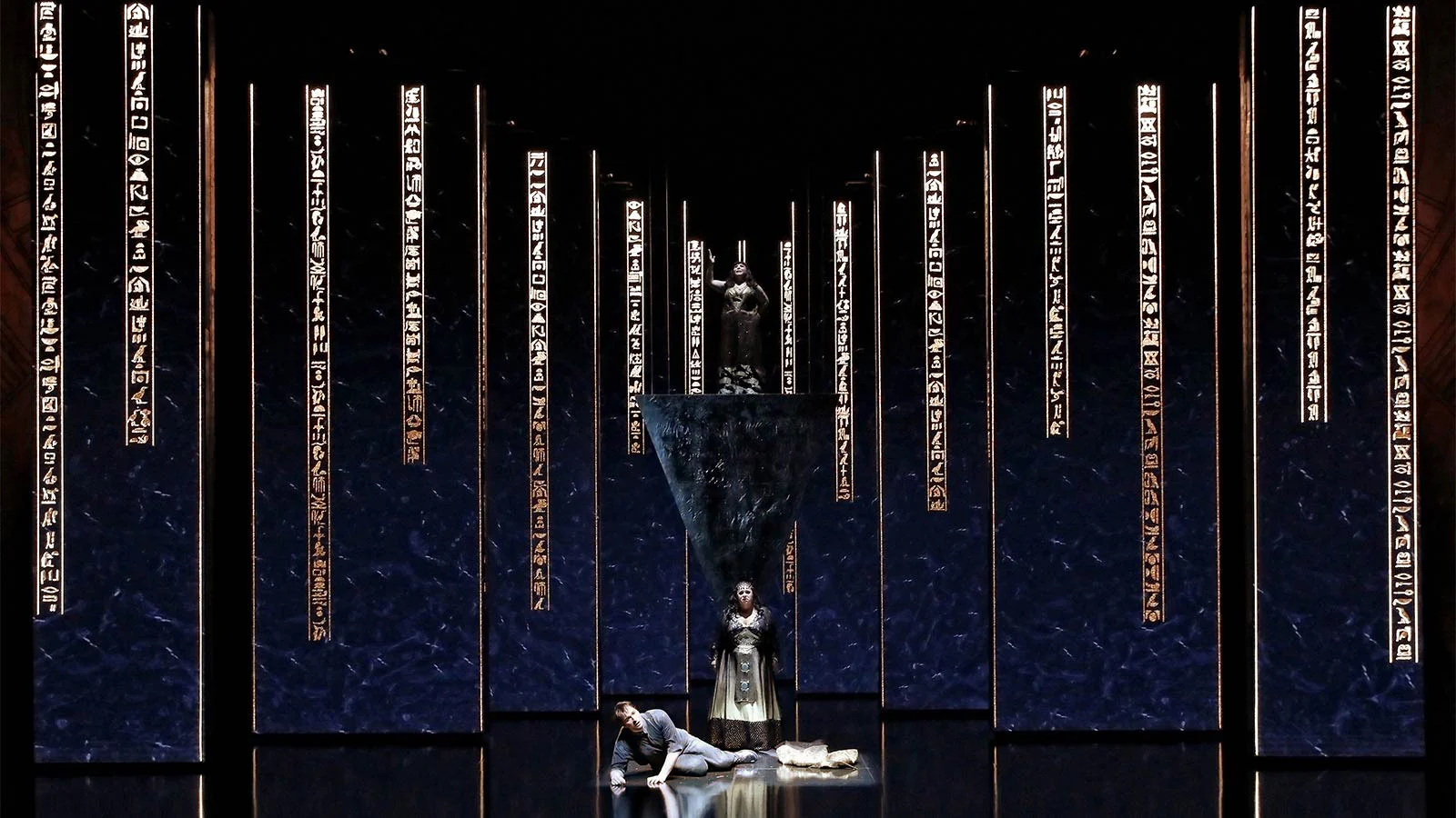 Schermi blu profondo con colonne di geroglifici dorati, una donna in piedi e un uomo a terra – Aida alla Sydney Opera House, Australia, 2018 – scenografia d'opera