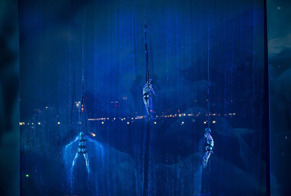 Dettaglio di tre artisti aerei appesi a funi davanti a una cortina d'acqua che cade, illuminati di blu – Cerimonia di apertura del Dubai Water Canal, Emirati Arabi Uniti, 2016 – progettazione di eventi
