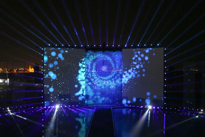 Palco di notte incorniciato da raggi di luce blu, con bolle proiettate e l'immagine di un vortice sugli schermi – Cerimonia di apertura del Dubai Water Canal, Emirati Arabi Uniti, 2016 – progettazione di eventi