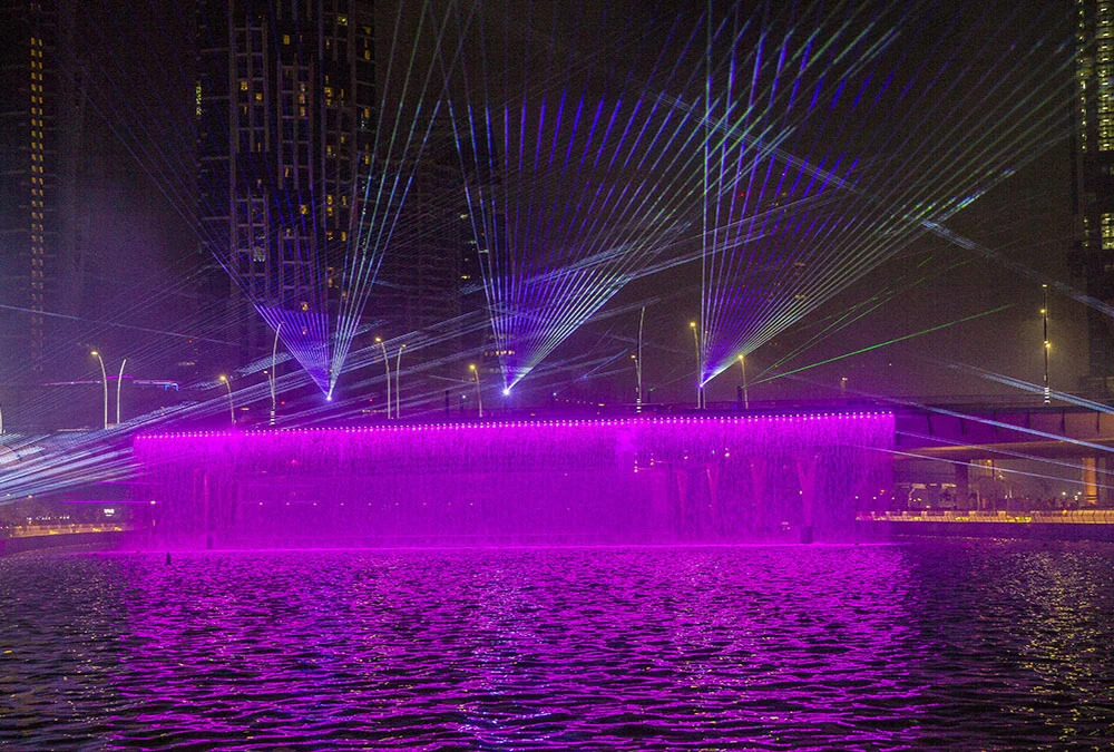 Parete d'acqua a cascata che brilla di magenta sotto ventagli di raggi laser, riflessa nel Dubai Water Canal di notte – Cerimonia di apertura del Dubai Water Canal, Emirati Arabi Uniti, 2016 – progettazione di eventi
