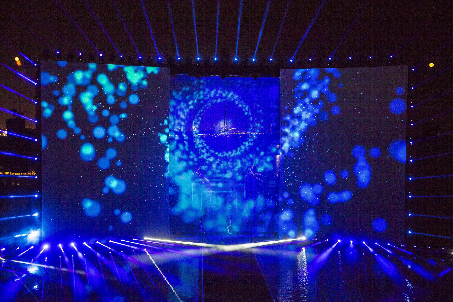 Vista frontale del palco con proiezioni di bolle blu sui tre schermi e un vortice centrale – Cerimonia di apertura del Dubai Water Canal, Emirati Arabi Uniti, 2016 – progettazione di eventi