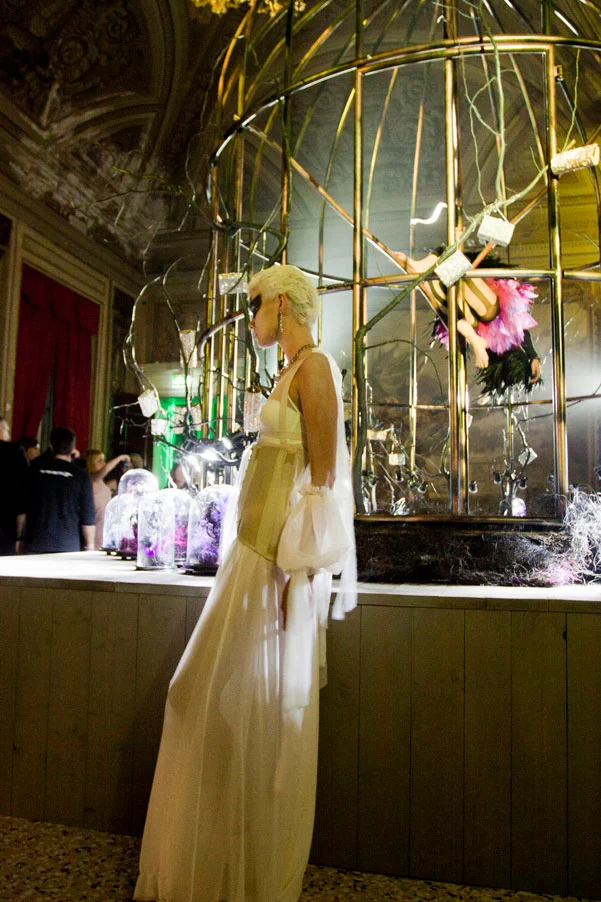 Modella in un lungo abito bianco in posa accanto alla gigantesca voliera dorata con le vetrine di gioielli – Swarovski Fashionation Party a Palazzo Reale, Milano, Italia, 2011 – progettazione di eventi