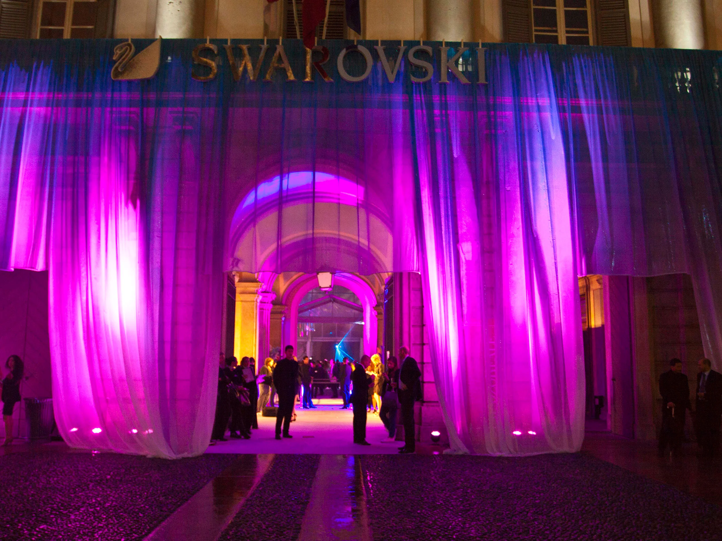 Ingresso di Palazzo Reale velato da tende leggere illuminate di magenta, con il logo Swarovski sopra il portale ad arco, di notte – Swarovski Fashionation Party a Palazzo Reale, Milano, Italia, 2011 – progettazione di eventi