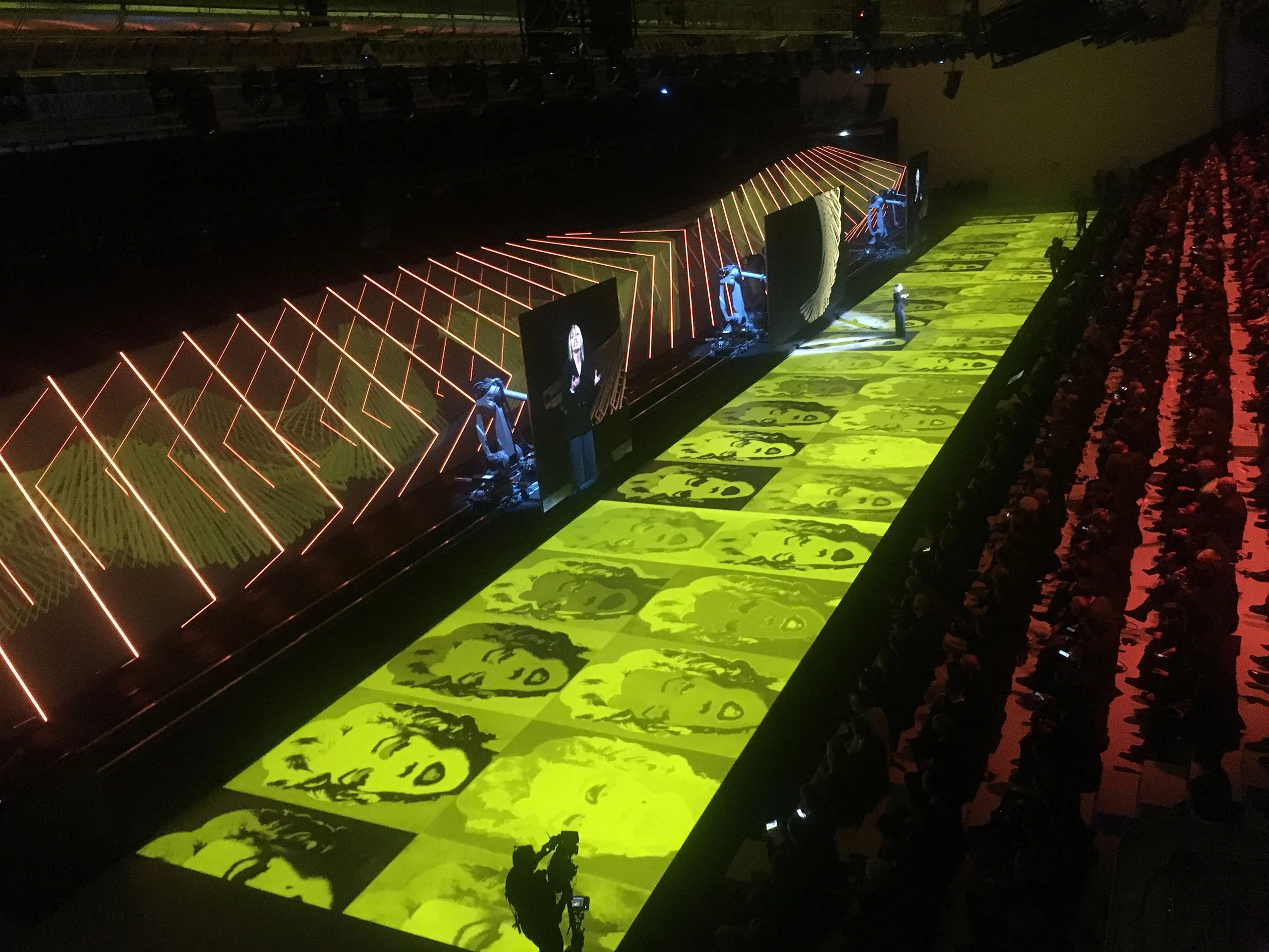 Lunga passerella illuminata di giallo con volti pop art proiettati, bracci robotici e cornici di luce arancioni accanto al pubblico – Anteprima mondiale Lamborghini Urus, Sant'Agata Bolognese, Italia, 2017 – progettazione di eventi