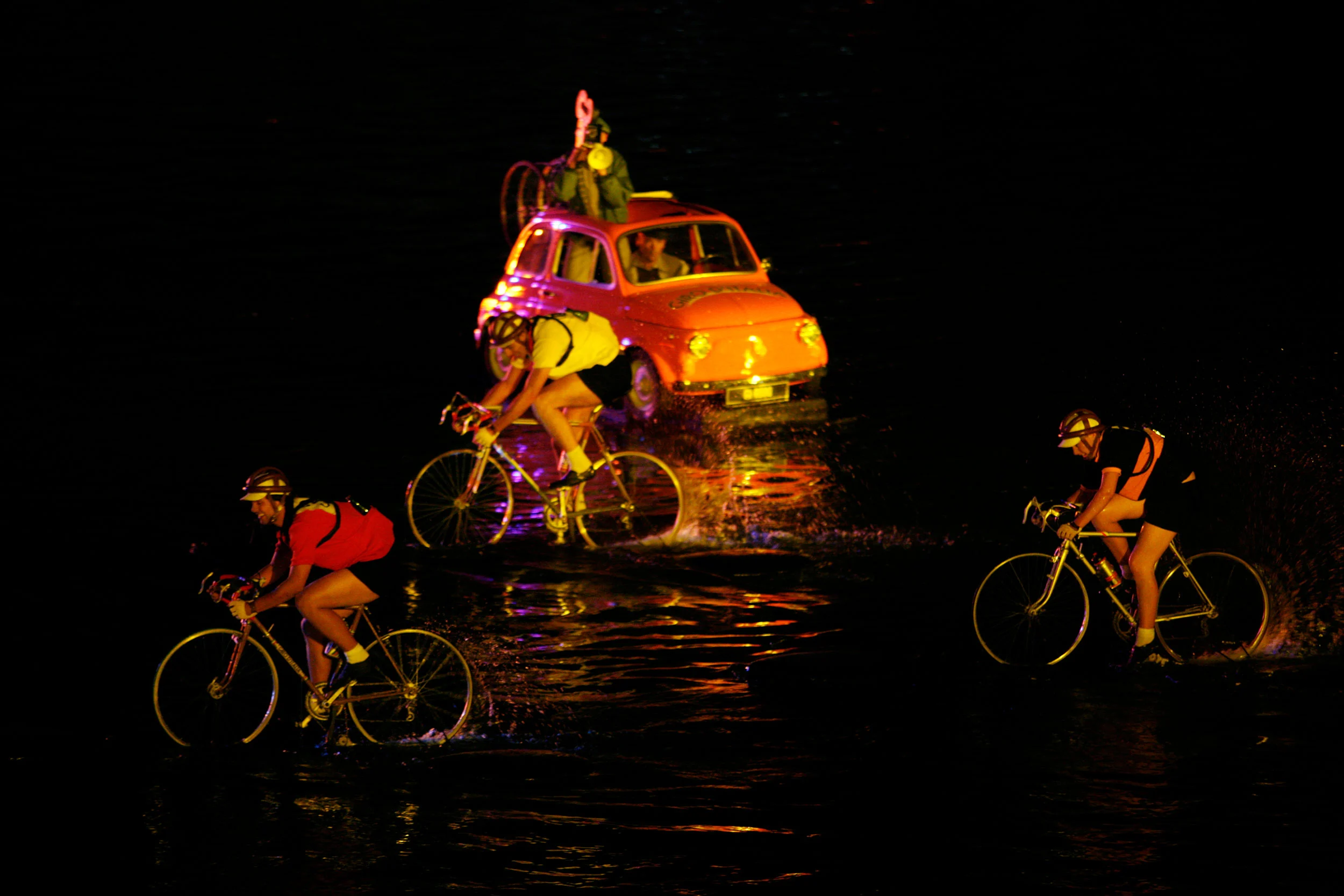Ciclisti in maglie da corsa d'epoca che attraversano l'acqua davanti a una Fiat 500 classica arancione di notte – Anteprima mondiale Fiat Welcome 500, Torino, Italia, 2007 – progettazione di eventi