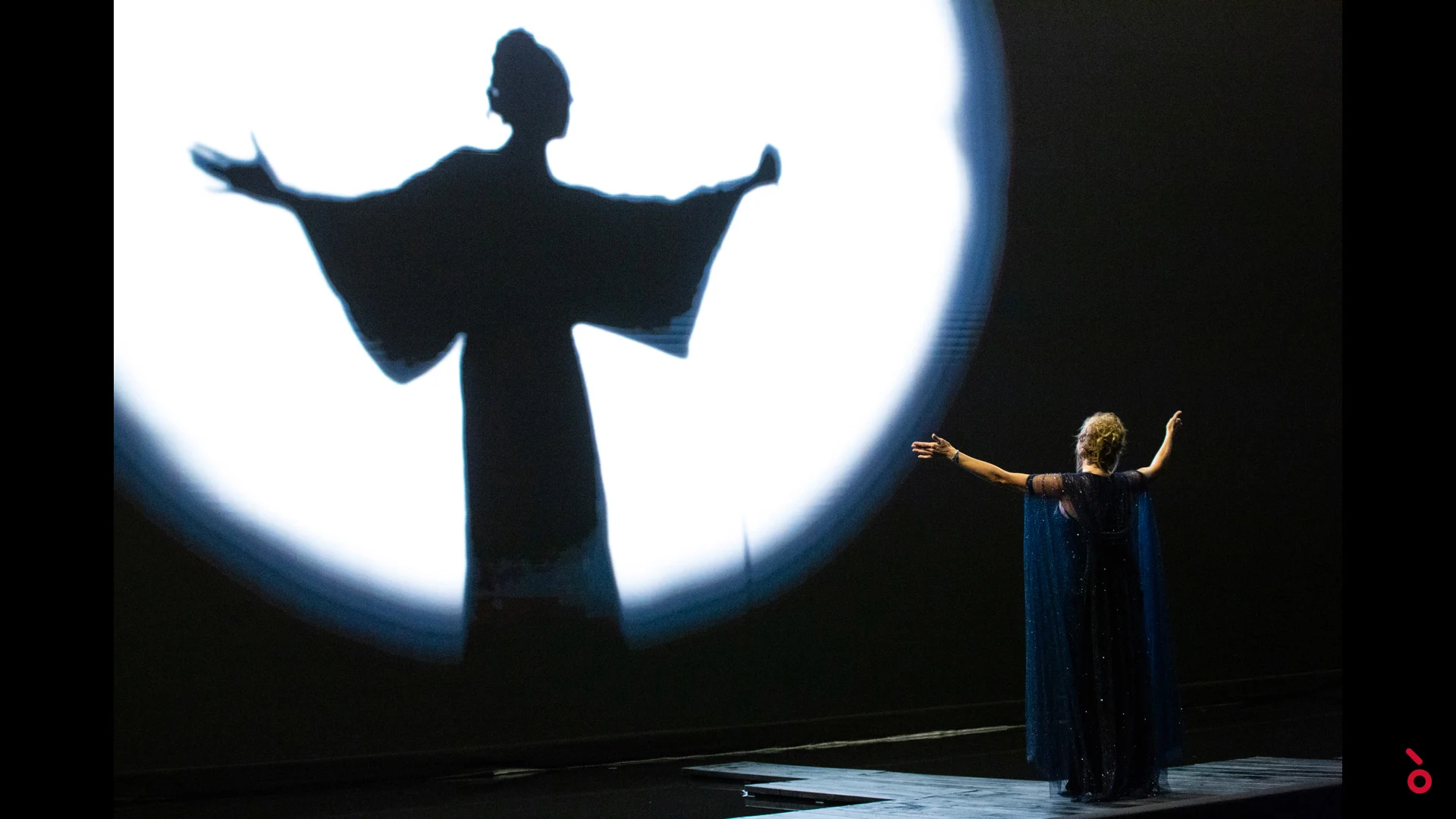 Cantante con mantello blu a braccia aperte di fronte alla sua ombra gigante in un cerchio di luce bianca – A Riveder le Stelle al Teatro alla Scala, Milano, Italia, 2020 – scenografia d'opera
