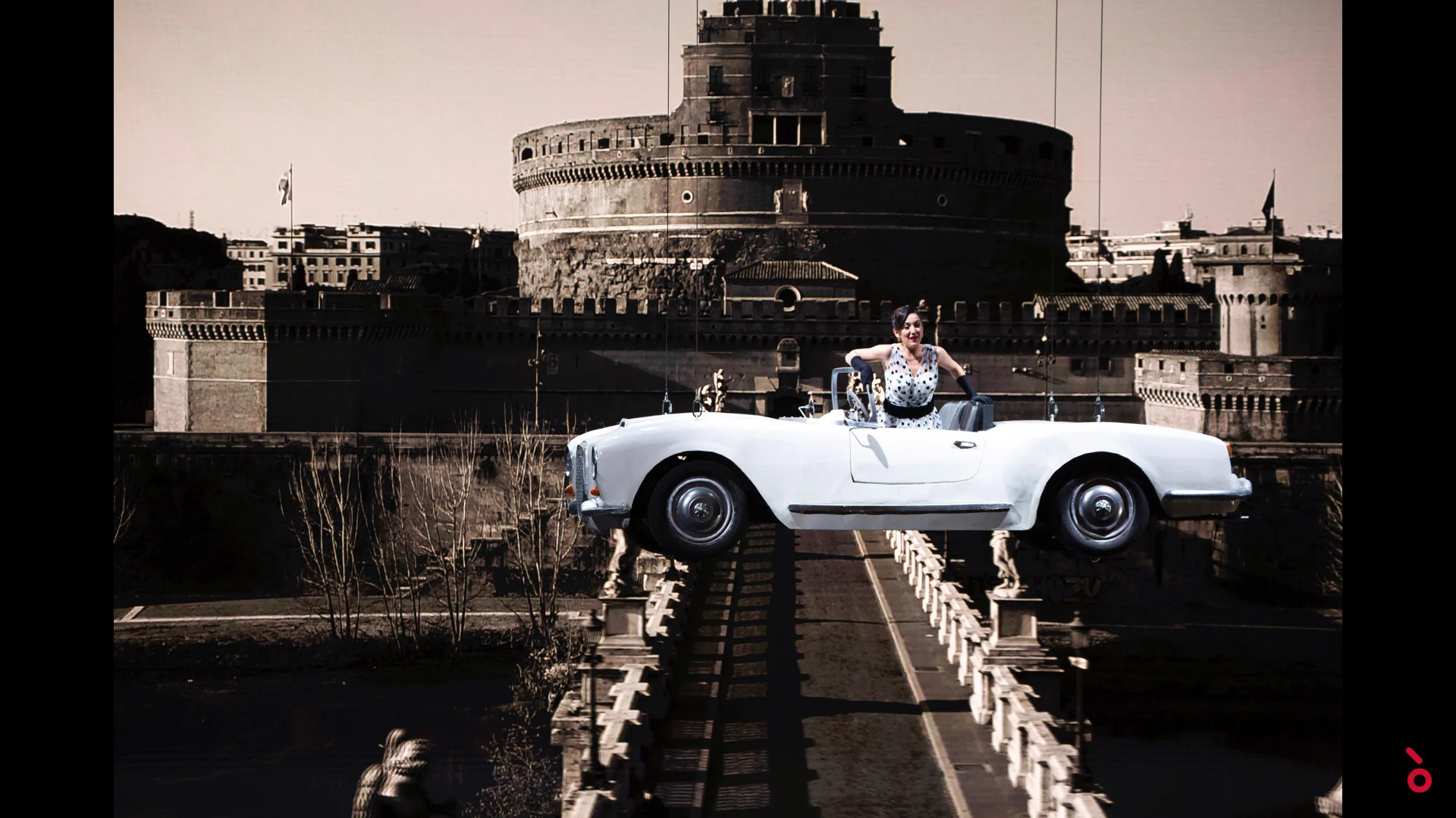Cantante in una decappottabile bianca che sembra sospesa su un ponte, Castel Sant'Angelo proiettato alle spalle – A Riveder le Stelle al Teatro alla Scala, Milano, Italia, 2020 – scenografia d'opera