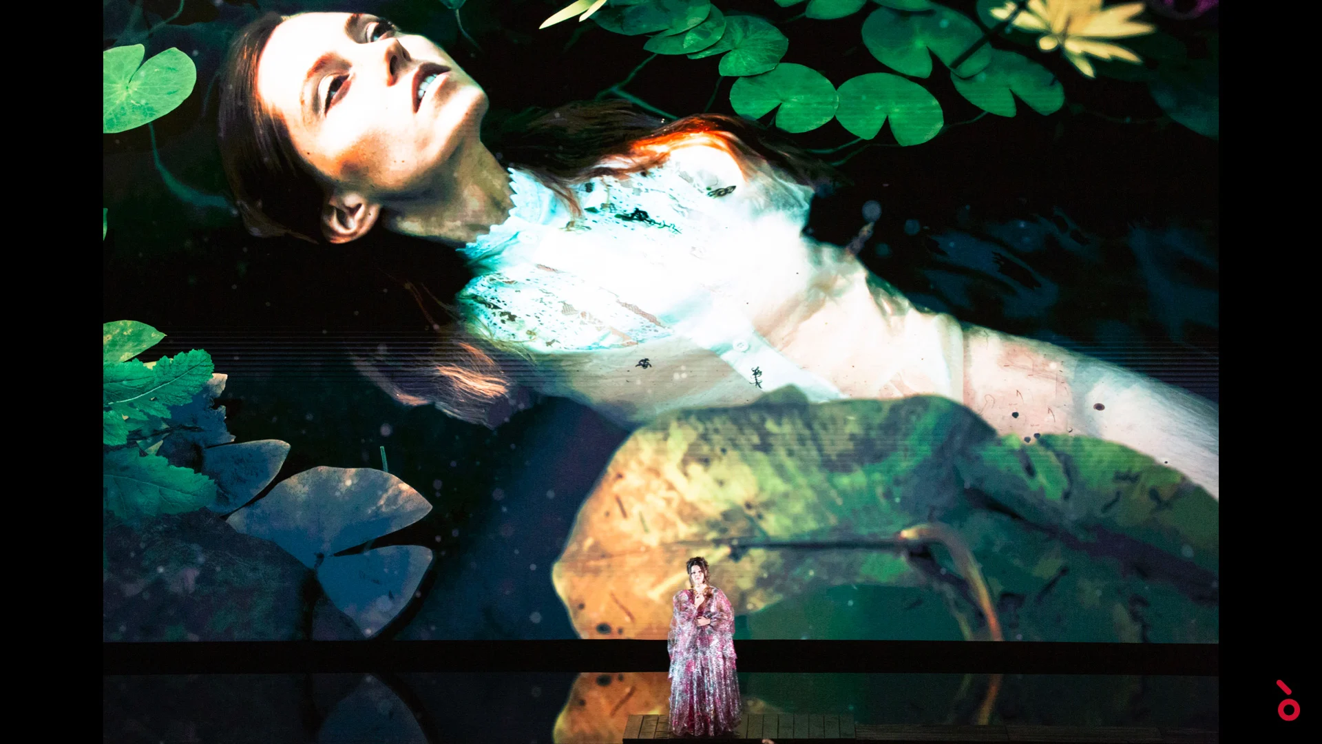 Cantante in abito lungo sovrastata dalla proiezione di una donna che galleggia tra le ninfee – A Riveder le Stelle al Teatro alla Scala, Milano, Italia, 2020 – scenografia d'opera