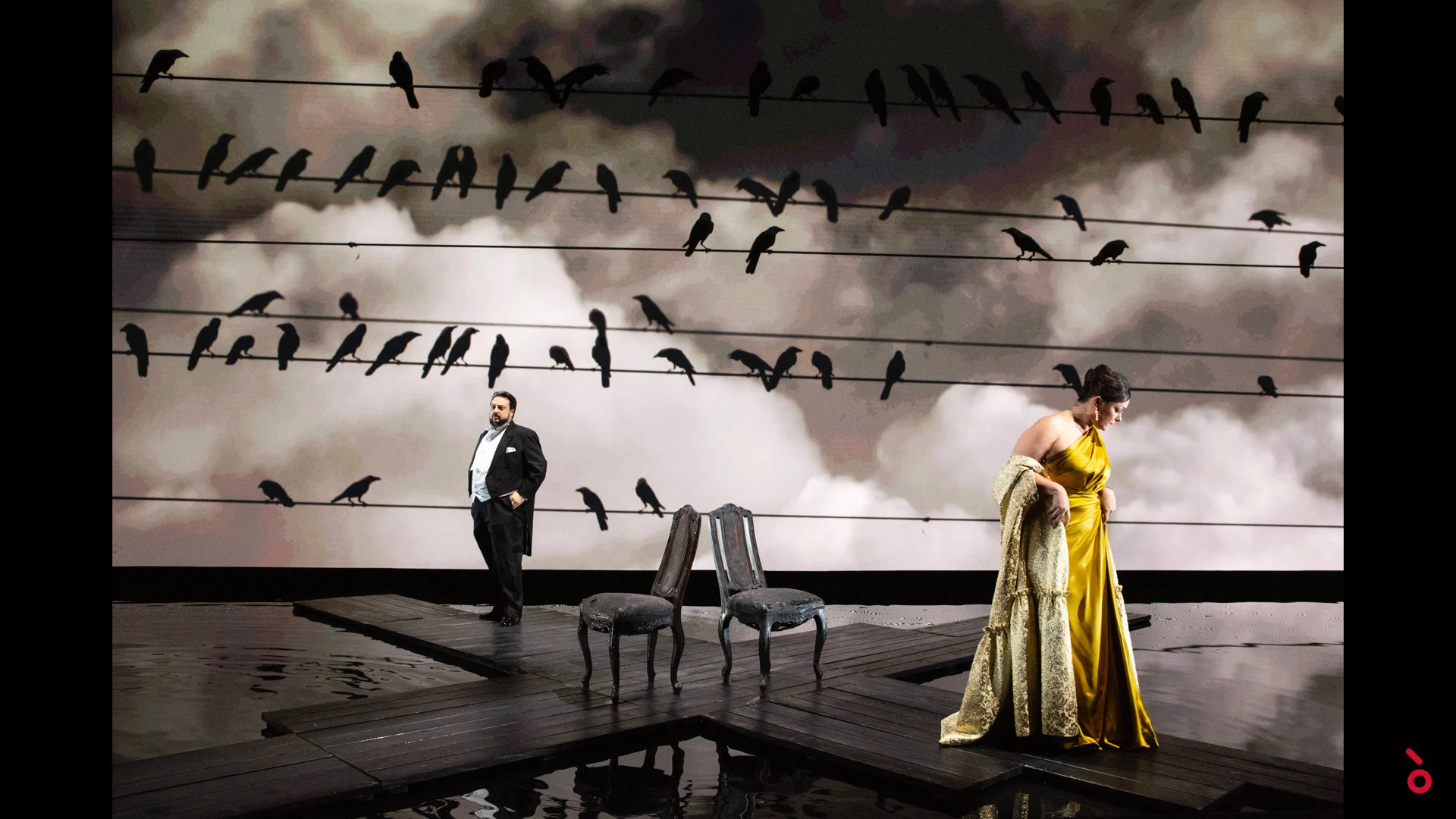 Due cantanti accanto a sedie vuote su un tavolato bagnato, corvi sui fili proiettati su un cielo nuvoloso – A Riveder le Stelle al Teatro alla Scala, Milano, Italia, 2020 – scenografia d'opera