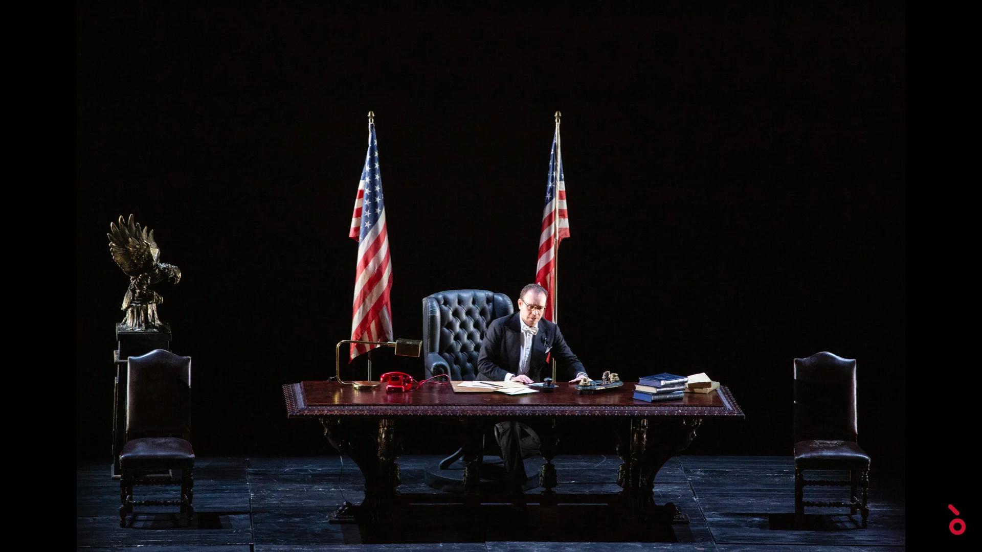 Attore seduto a una grande scrivania tra due bandiere americane, una scultura d'aquila a sinistra – A Riveder le Stelle al Teatro alla Scala, Milano, Italia, 2020 – scenografia d'opera