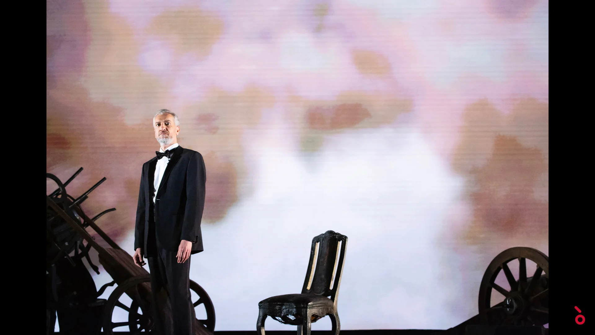 Cantante in smoking accanto a una sedia vuota e vecchie ruote di carro, morbide nuvole rosa proiettate alle spalle – A Riveder le Stelle al Teatro alla Scala, Milano, Italia, 2020 – scenografia d'opera