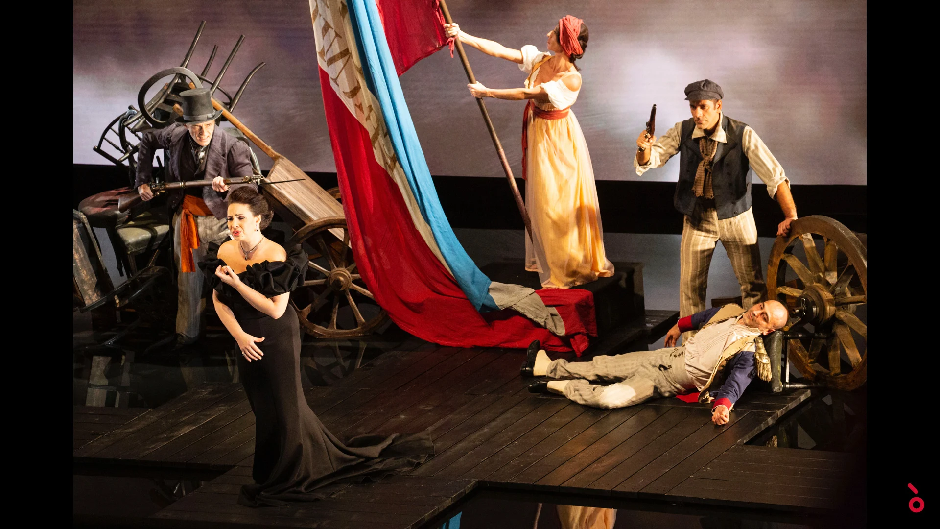 Cantante in abito nero davanti a una scena di barricata con un grande tricolore, ruote di carro e attori – A Riveder le Stelle al Teatro alla Scala, Milano, Italia, 2020 – scenografia d'opera