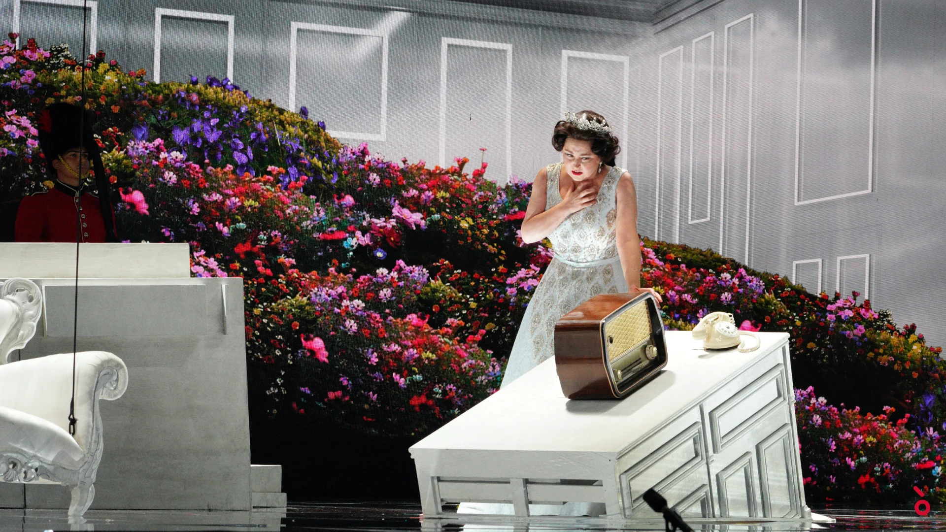 La regina in abito bianco accanto a una vecchia radio su un cassettone bianco, davanti a una parete di fiori colorati – Elisabetta, Regina d'Inghilterra al Rossini Opera Festival, Pesaro, Italia, 2021 – scenografia d'opera