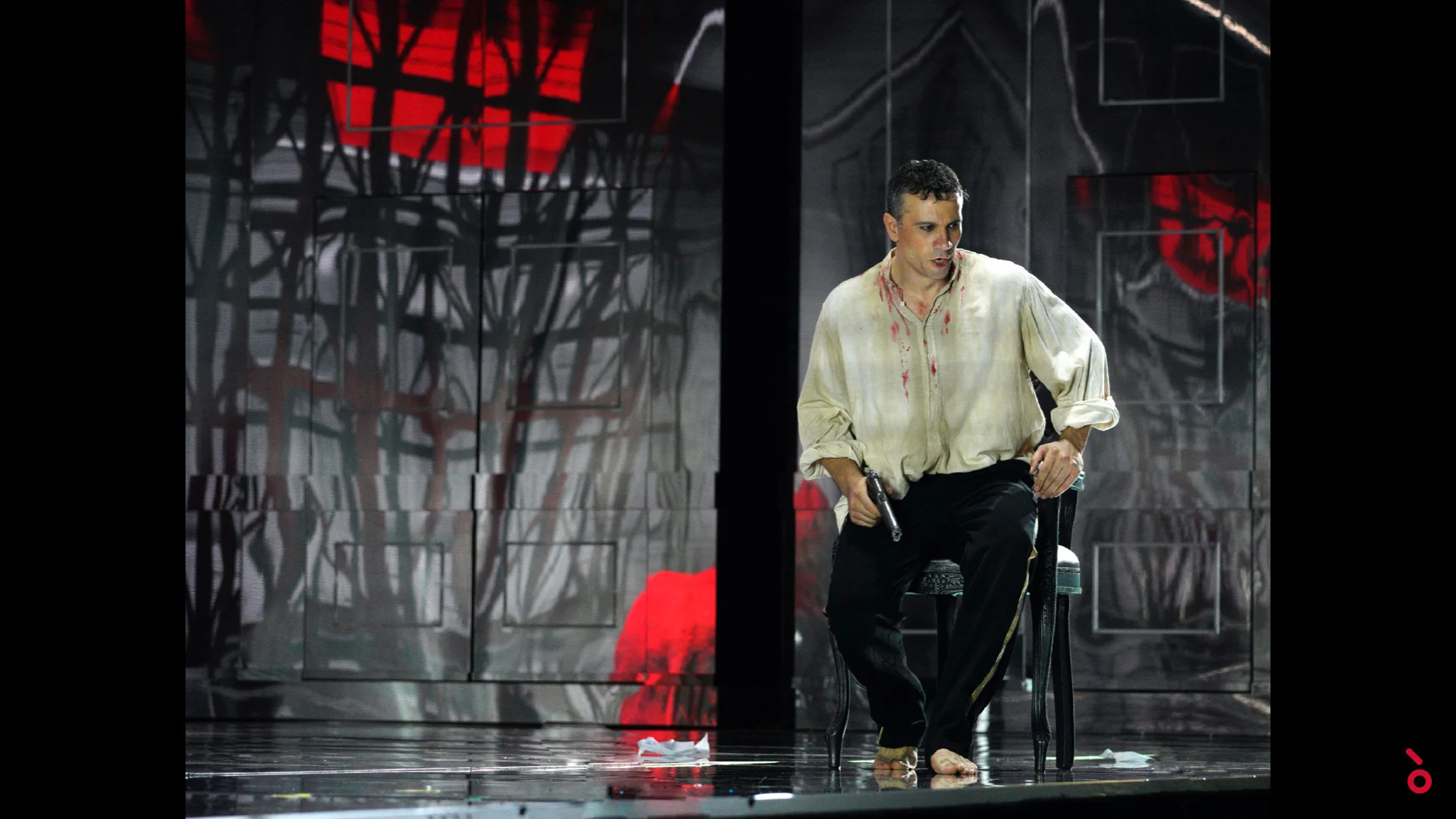Cantante in camicia macchiata di sangue seduto su una sedia, alberi spogli e luce rossa proiettati alle spalle – Elisabetta, Regina d'Inghilterra al Rossini Opera Festival, Pesaro, Italia, 2021 – scenografia d'opera