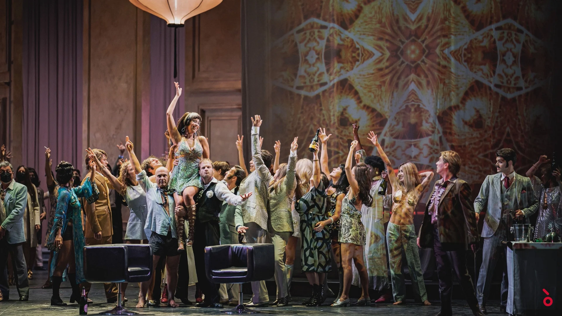 Scena di festa con il coro in costumi anni Sessanta a braccia alzate sotto una lanterna di carta, proiezione caleidoscopica alle spalle – La Traviata al Maggio Musicale Fiorentino, Firenze, Italia, 2022 – scenografia d'opera