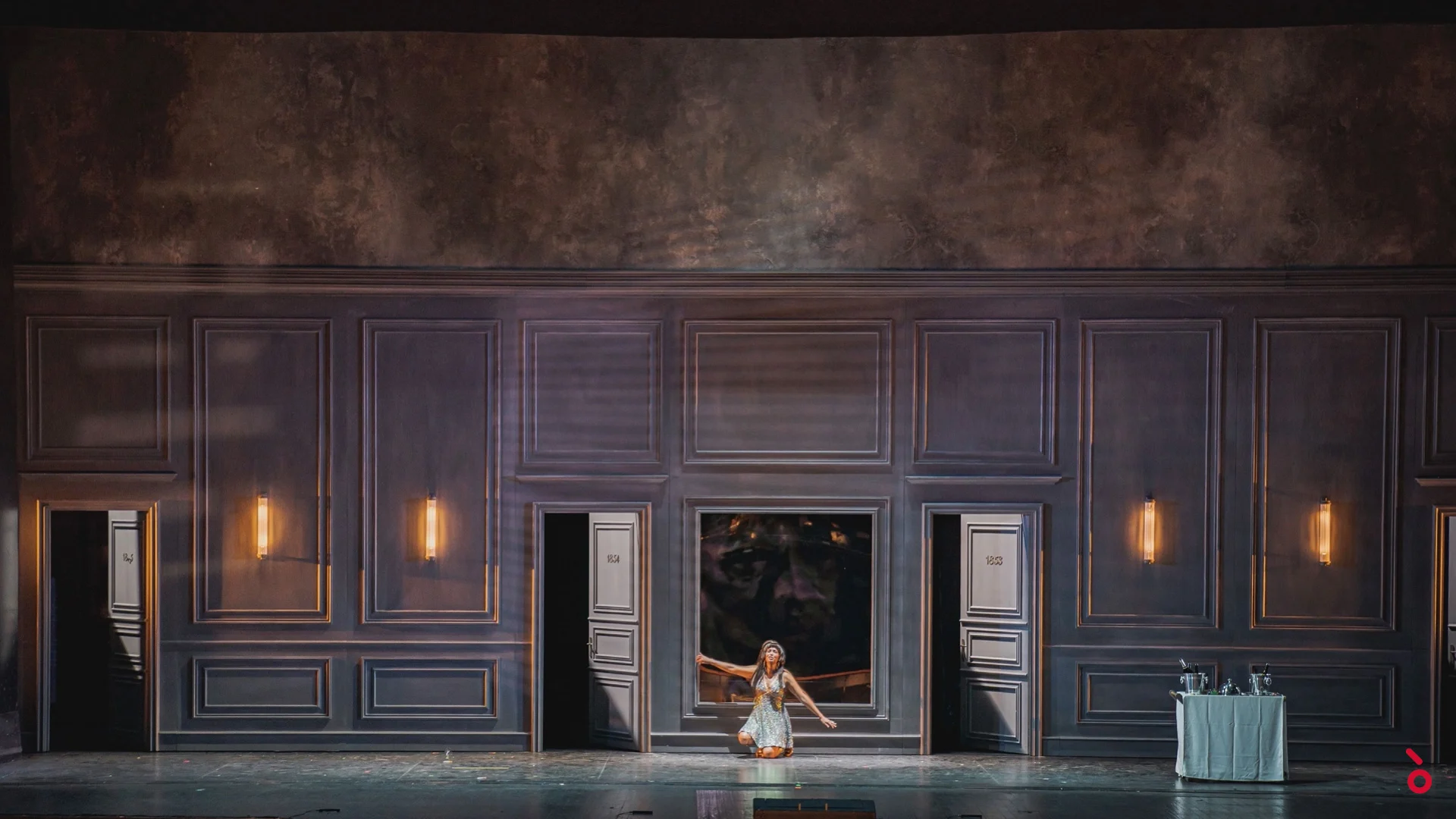 Vista d'insieme della scenografia a pannelli grigi con porte numerate e applique, una donna inginocchiata al centro – La Traviata al Maggio Musicale Fiorentino, Firenze, Italia, 2022 – scenografia d'opera