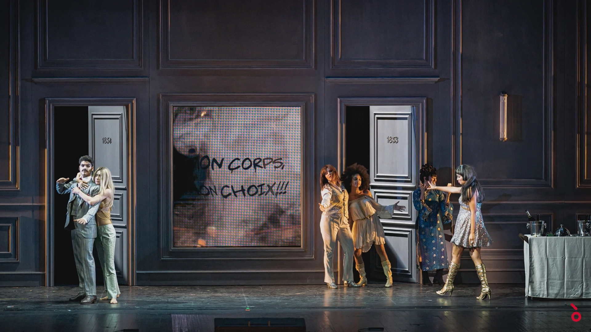 Coppie che escono da porte numerate d'albergo, lo slogan "Mon corps, mon choix" proiettato su uno schermo – La Traviata al Maggio Musicale Fiorentino, Firenze, Italia, 2022 – scenografia d'opera