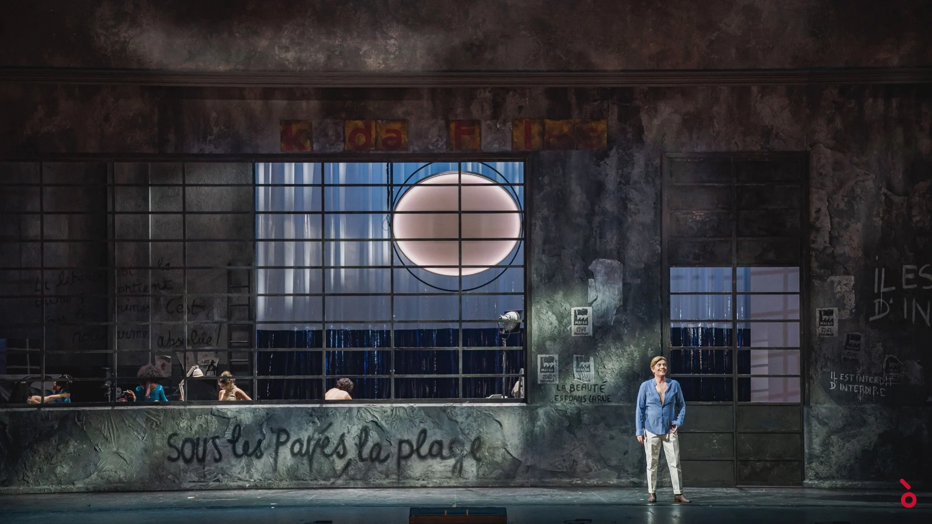 Muro consunto con grandi finestre e la scritta "Sous les pavés la plage", una cantante sola davanti – La Traviata al Maggio Musicale Fiorentino, Firenze, Italia, 2022 – scenografia d'opera