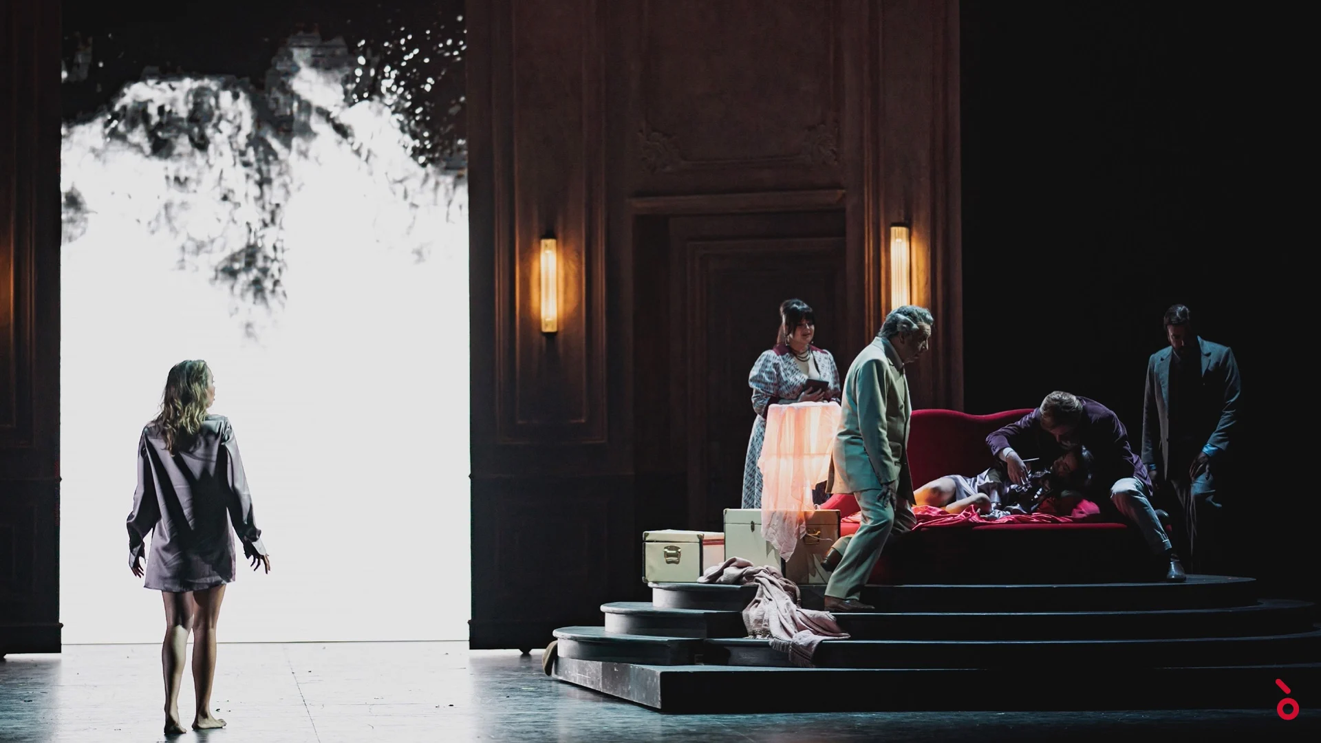 Una donna in camicia davanti a una porta bianca luminosa mentre altri si raccolgono attorno a un divano rosso su pedane a gradoni – La Traviata al Maggio Musicale Fiorentino, Firenze, Italia, 2022 – scenografia d'opera