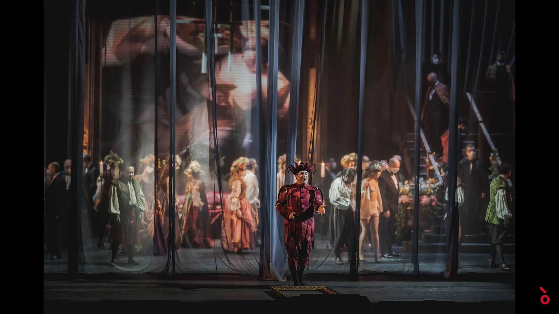 Buffone in costume rosso davanti a tende velate, cortigiani alle spalle e un grande volto proiettato – Rigoletto al Maggio Musicale Fiorentino, Firenze, Italia, 2021 – scenografia d'opera