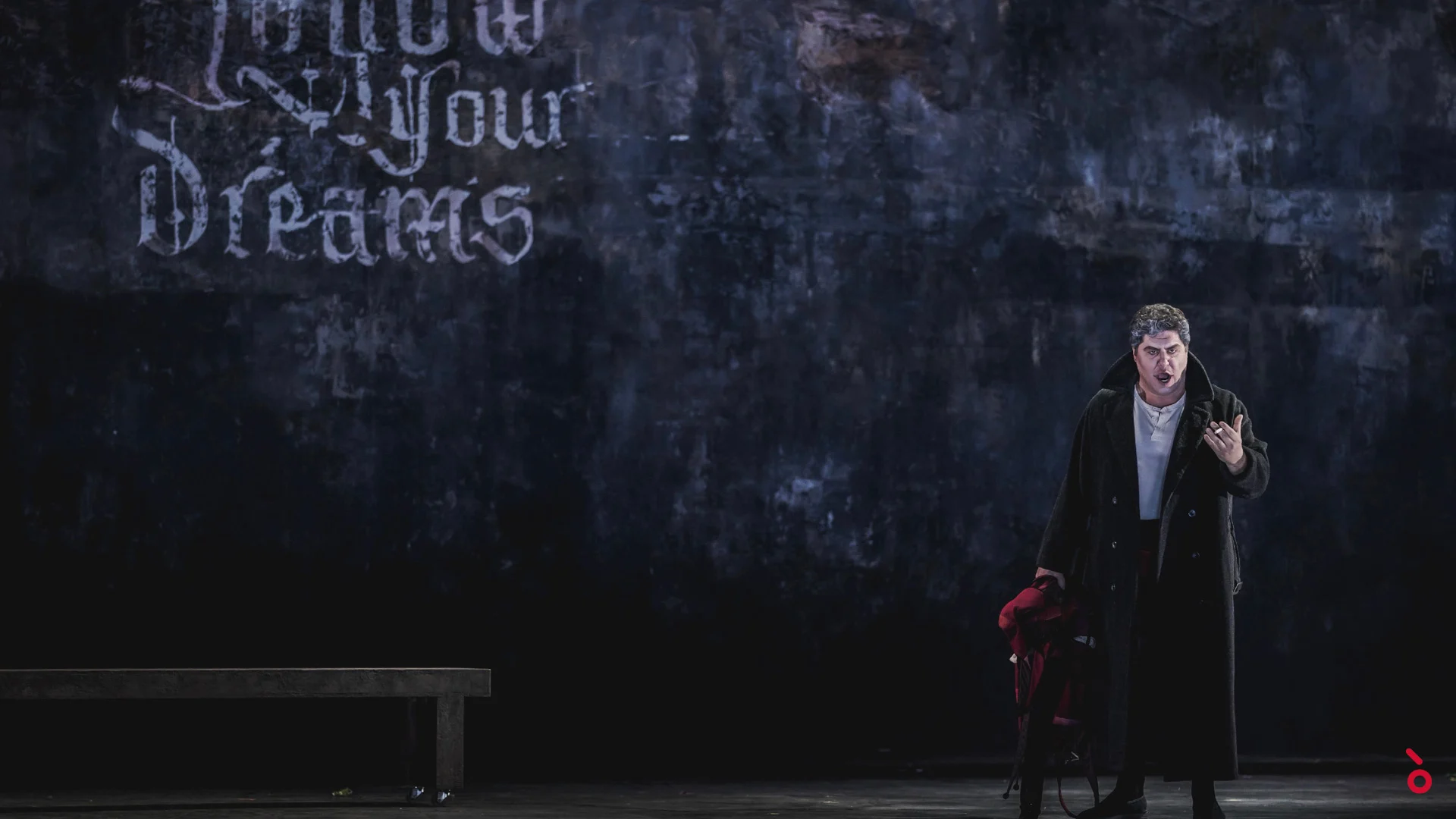 Cantante in un lungo cappotto nero davanti a un muro scuro con il graffito Follow Your Dreams, una panchina a sinistra – Rigoletto al Maggio Musicale Fiorentino, Firenze, Italia, 2021 – scenografia d'opera