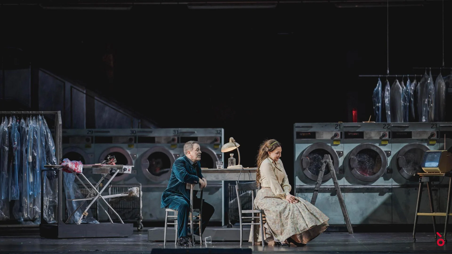 Due cantanti seduti a un tavolo nella scenografia della lavanderia, lavatrici e abiti impacchettati alle spalle – Rigoletto al Maggio Musicale Fiorentino, Firenze, Italia, 2021 – scenografia d'opera