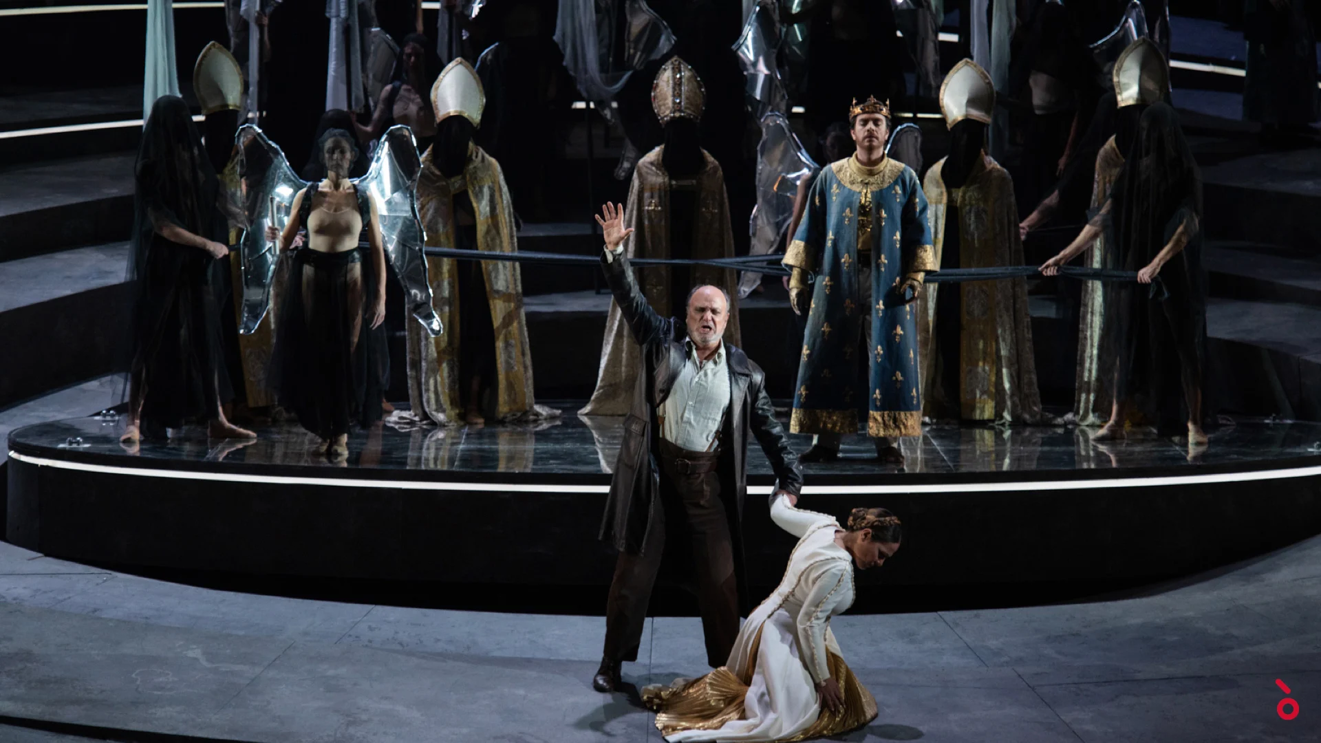 Cantante in cappotto di pelle sul palco circolare, una donna inginocchiata ai suoi piedi, il coro con le mitre alle spalle – Giovanna d'Arco al Teatro alla Scala, Milano, Italia, 2021 – scenografia d'opera