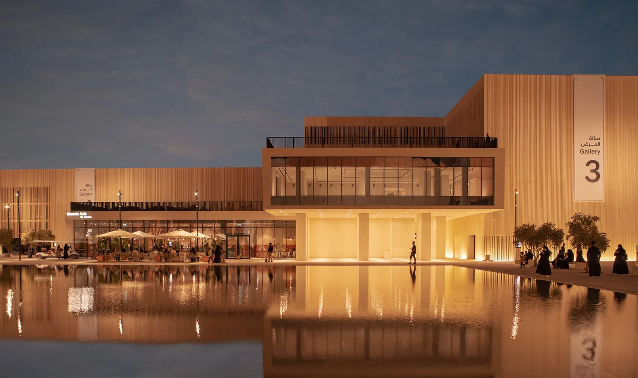 Padiglione Gallery 3 al tramonto, la facciata a listelli illuminata da luce calda e il piano superiore vetrato riflessi in una lunga vasca – Biennale delle Arti Islamiche, Jeddah, Arabia Saudita, 2023 – architettura