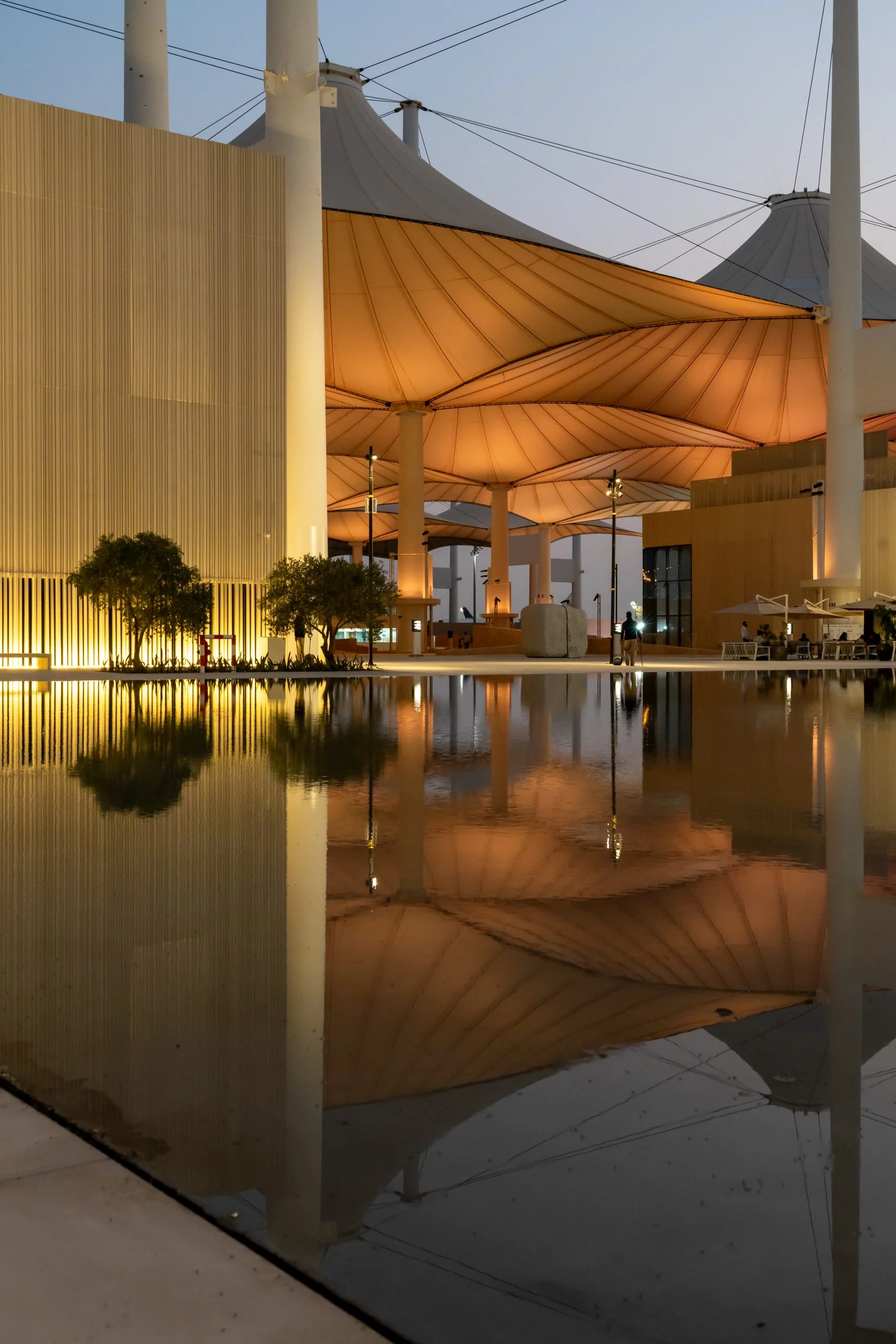 Coperture in tessuto dell'Hajj Terminal illuminate al tramonto accanto a un padiglione a listelli, riflesse nell'acqua ferma – Biennale delle Arti Islamiche, Jeddah, Arabia Saudita, 2023 – architettura