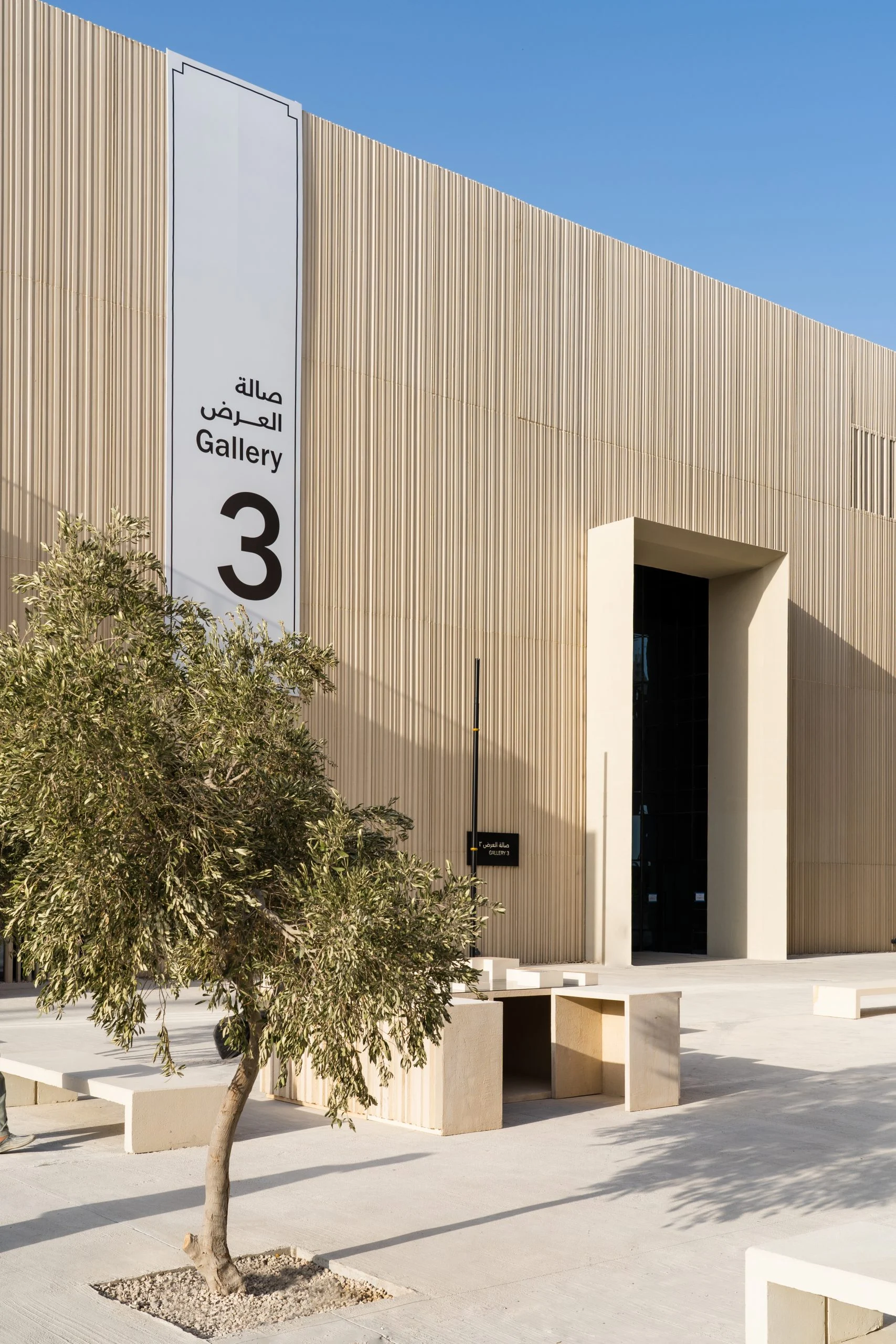 Facciata beige a listelli della Gallery 3 con segnaletica bilingue, un ulivo e panche in pietra al sole – Biennale delle Arti Islamiche, Jeddah, Arabia Saudita, 2023 – architettura