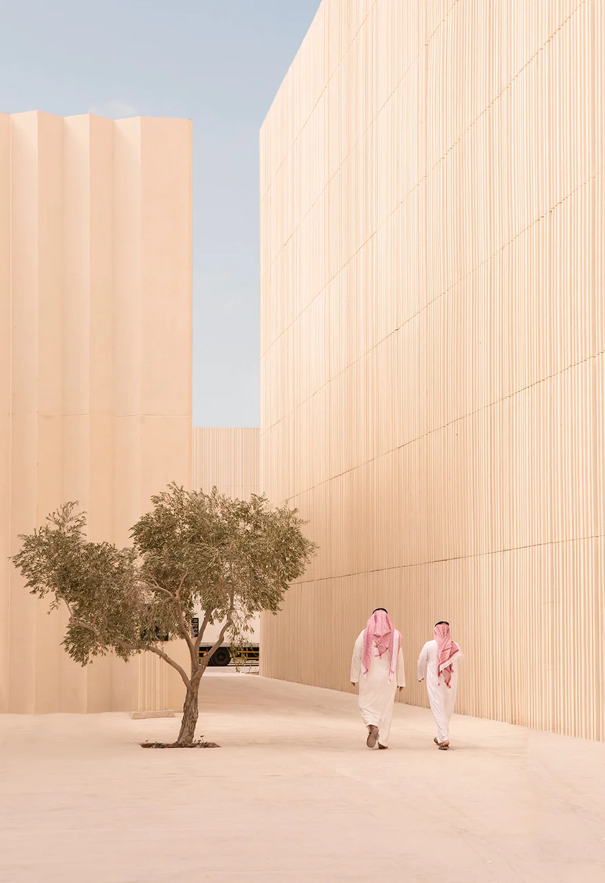 Due visitatori in thobe bianche che passano accanto a un ulivo tra alte pareti chiare a listelli del padiglione – Biennale delle Arti Islamiche, Jeddah, Arabia Saudita, 2023 – architettura