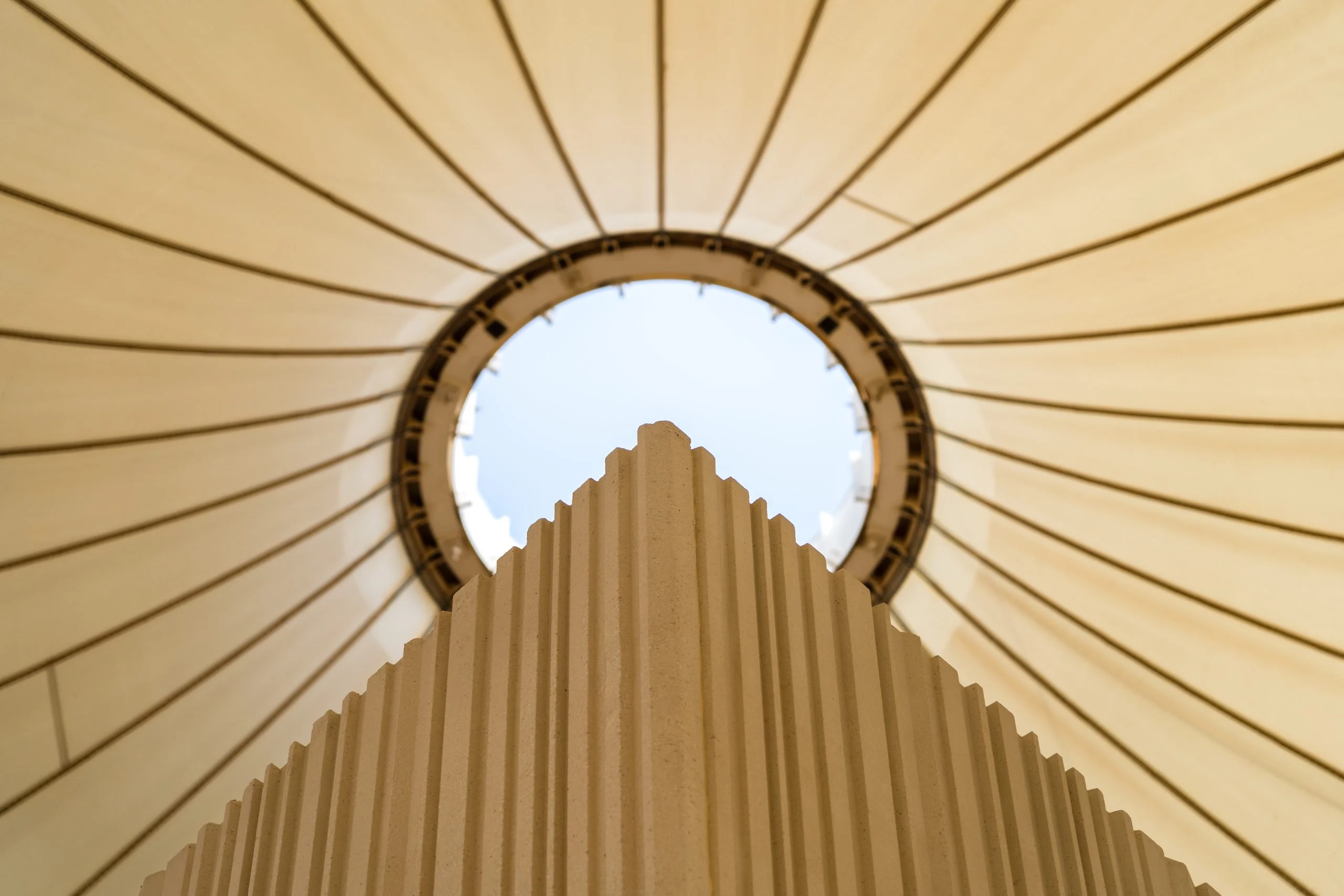 Vista dal basso di una colonna scultorea nervata che sale verso l'oculo circolare di una copertura a tenda in tessuto – Biennale delle Arti Islamiche, Jeddah, Arabia Saudita, 2023 – architettura