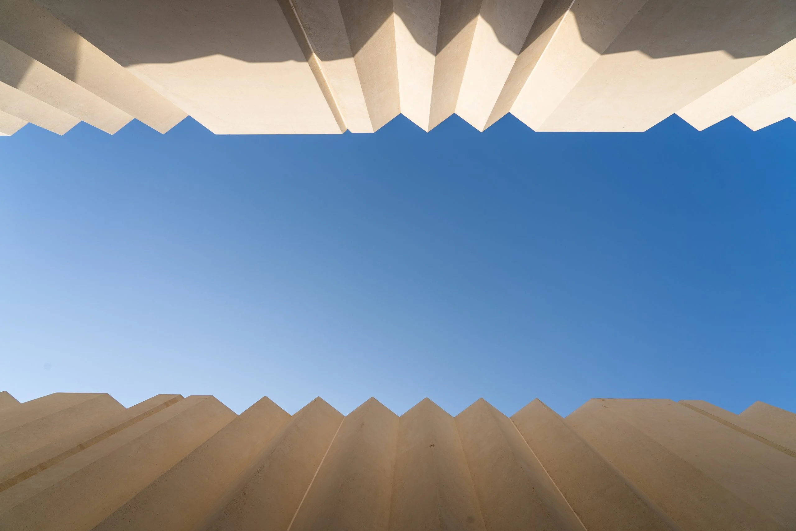 Vista dal basso del cielo blu incorniciato dai bordi a zigzag di due pareti chiare piegate – Biennale delle Arti Islamiche, Jeddah, Arabia Saudita, 2023 – architettura