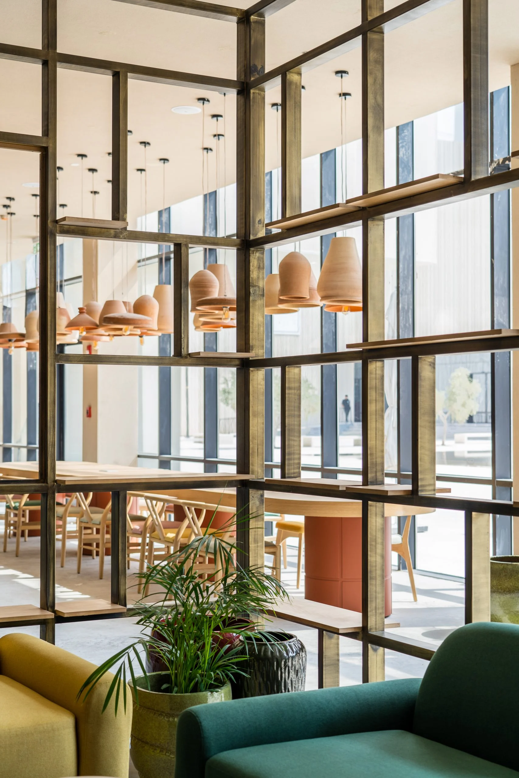 Scaffalatura a griglia in ottone che divide il caffè, con lampade in terracotta, palme in vaso e poltrone verdi – Biennale delle Arti Islamiche, Jeddah, Arabia Saudita, 2023 – architettura