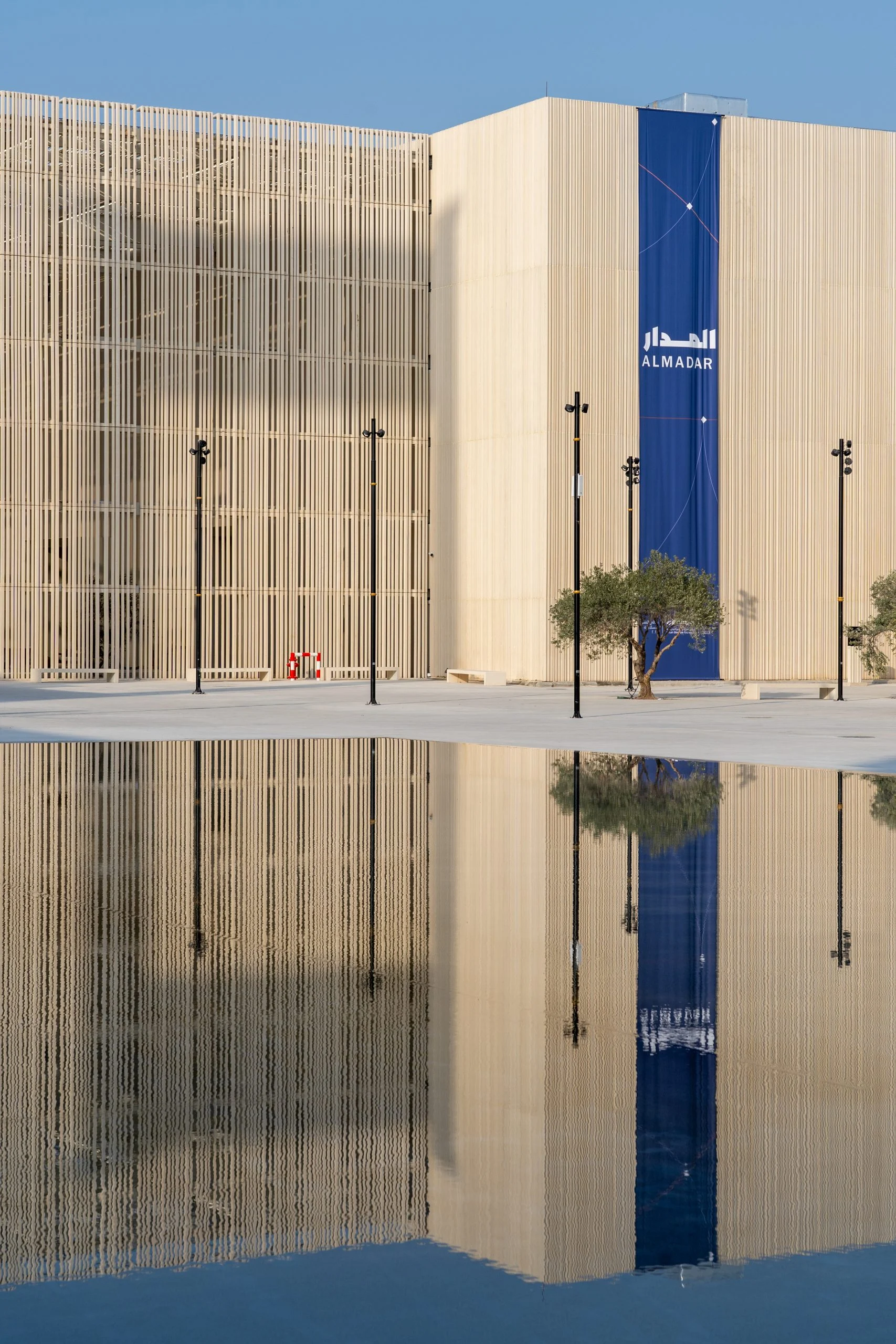 Facciata a listelli di un padiglione con uno striscione blu AlMadar, riflessa in una vasca a specchio sotto un cielo sereno – Biennale delle Arti Islamiche, Jeddah, Arabia Saudita, 2023 – architettura